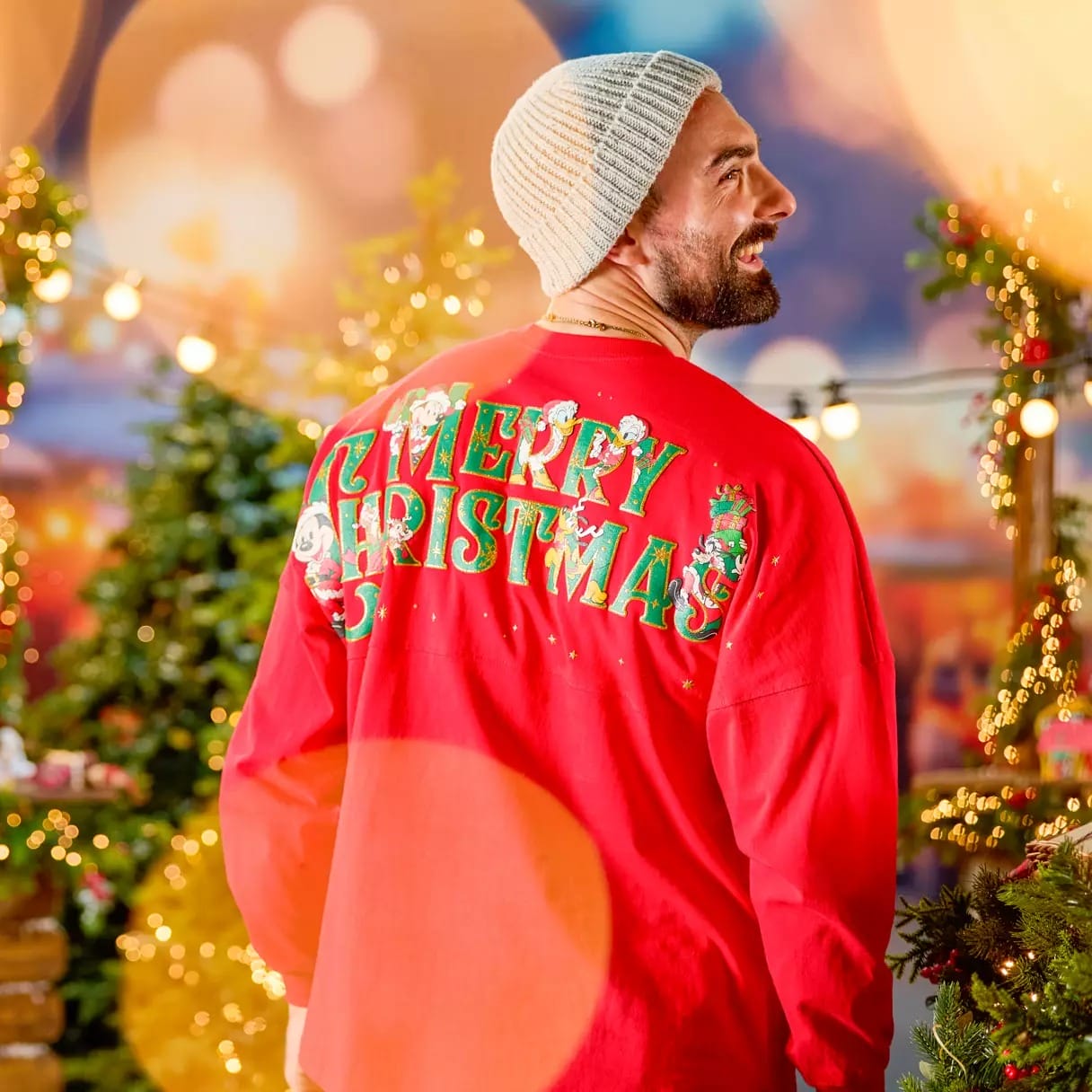 A smiling man in a red "Merry Christmas" shirt and knit beanie stands by decorated trees and festive lights.