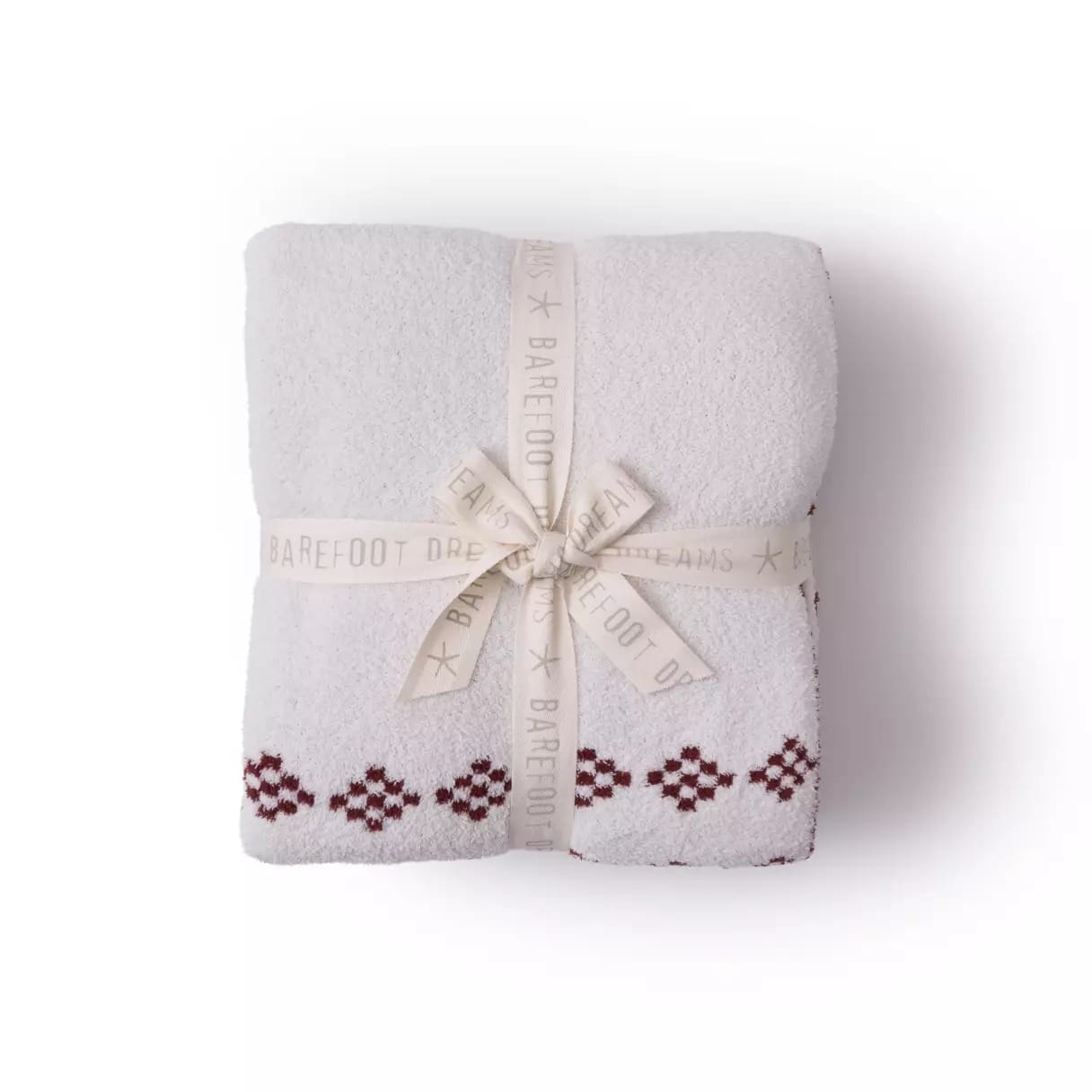 Folded white blanket with red geometric trim, tied with a cream ribbon that reads "BAREFOOT DREAMS" in gray text.