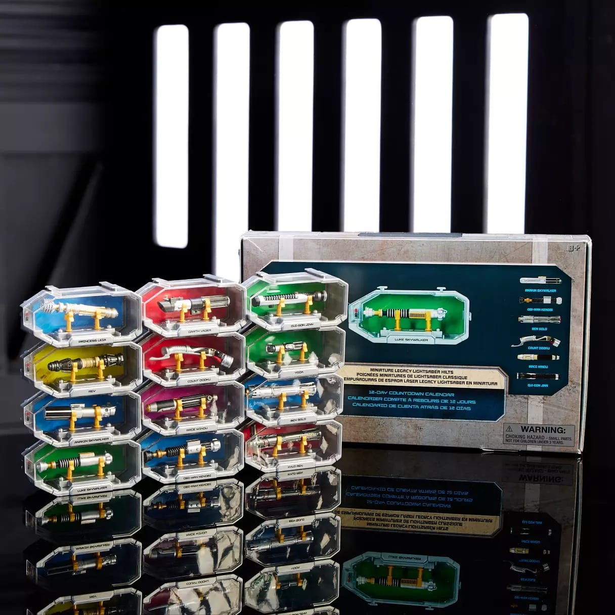 Six colored Star Wars lightsaber hilts in display cases, with packaging, like those sold at Disney Parks, on a reflective surface.