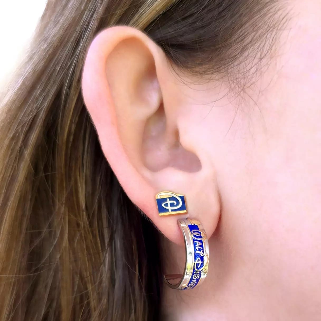Close-up of an ear wearing a blue and gold earring with a "D" design, inspired by classic Disney iconography and decorative details.