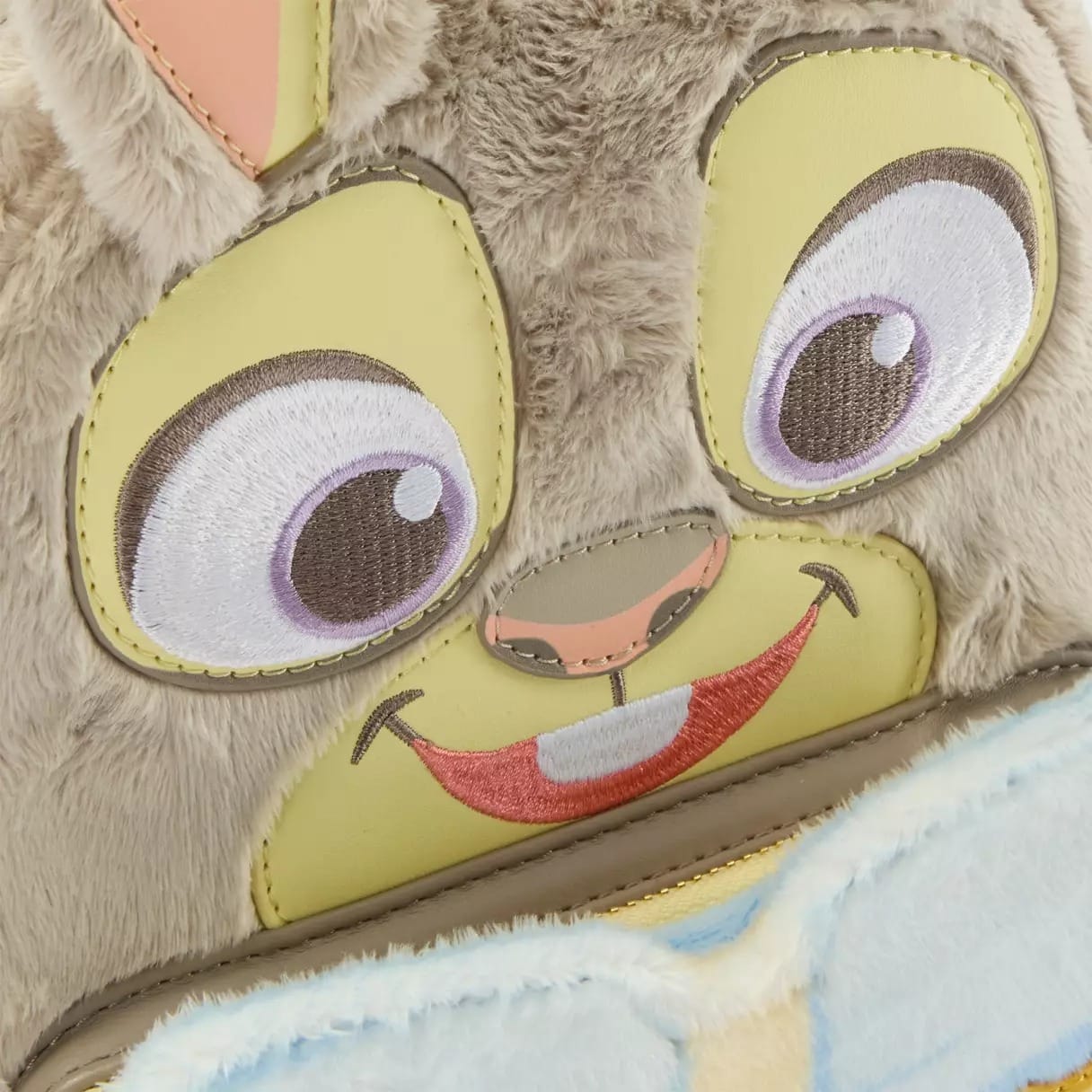 Close-up of a plush animal face, reminiscent of a Disney park character, with big embroidered eyes, pink nose, and bucktooth smile.