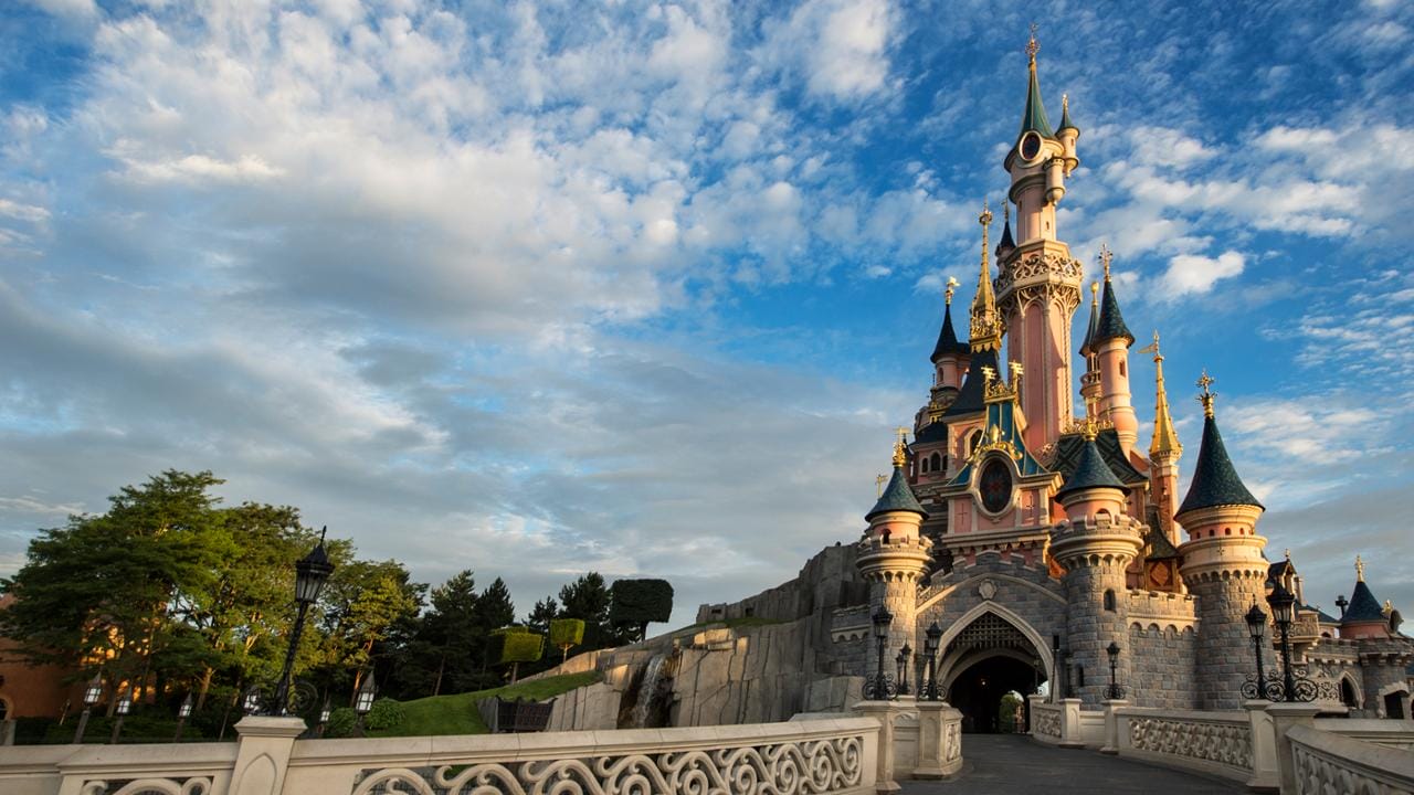 A fairytale castle with pink walls and blue turrets, resembling Sleeping Beauty Castle at Disneyland, beneath a blue sky with clouds.