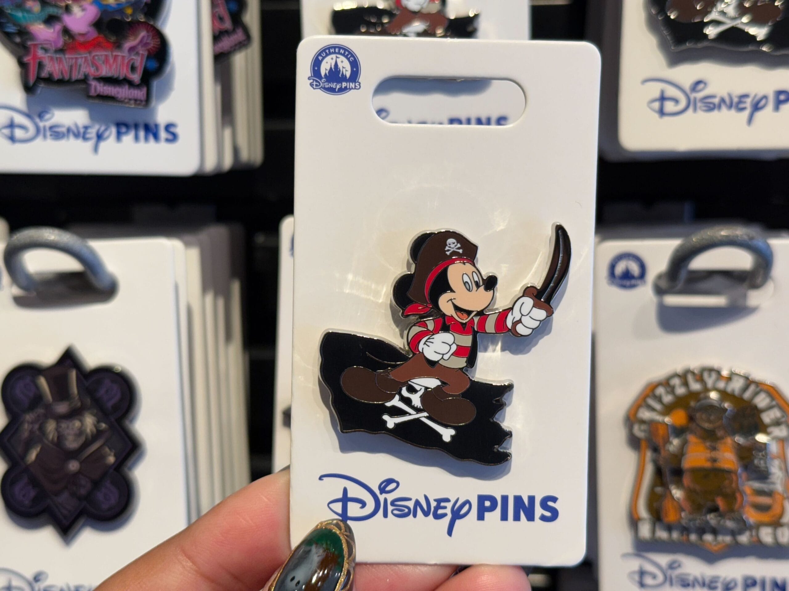A hand holds a Disney pin of pirate Mickey Mouse with a sword, with more Disney pins—like those sold in parks—seen in the background.