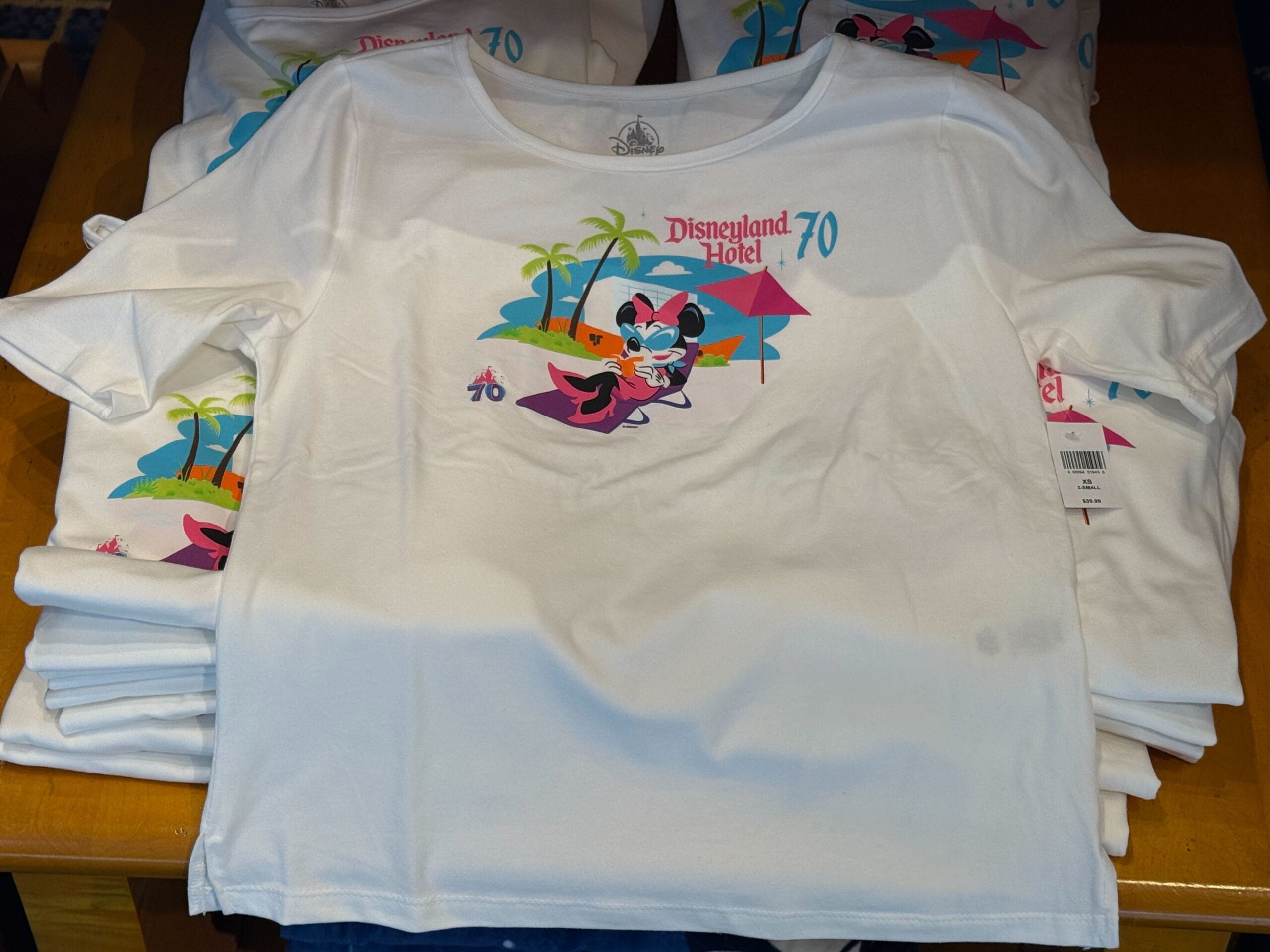 White t-shirt with Minnie Mouse, palm trees, and Disneyland Hotel 70th Anniversary logo, stacked on a display table.