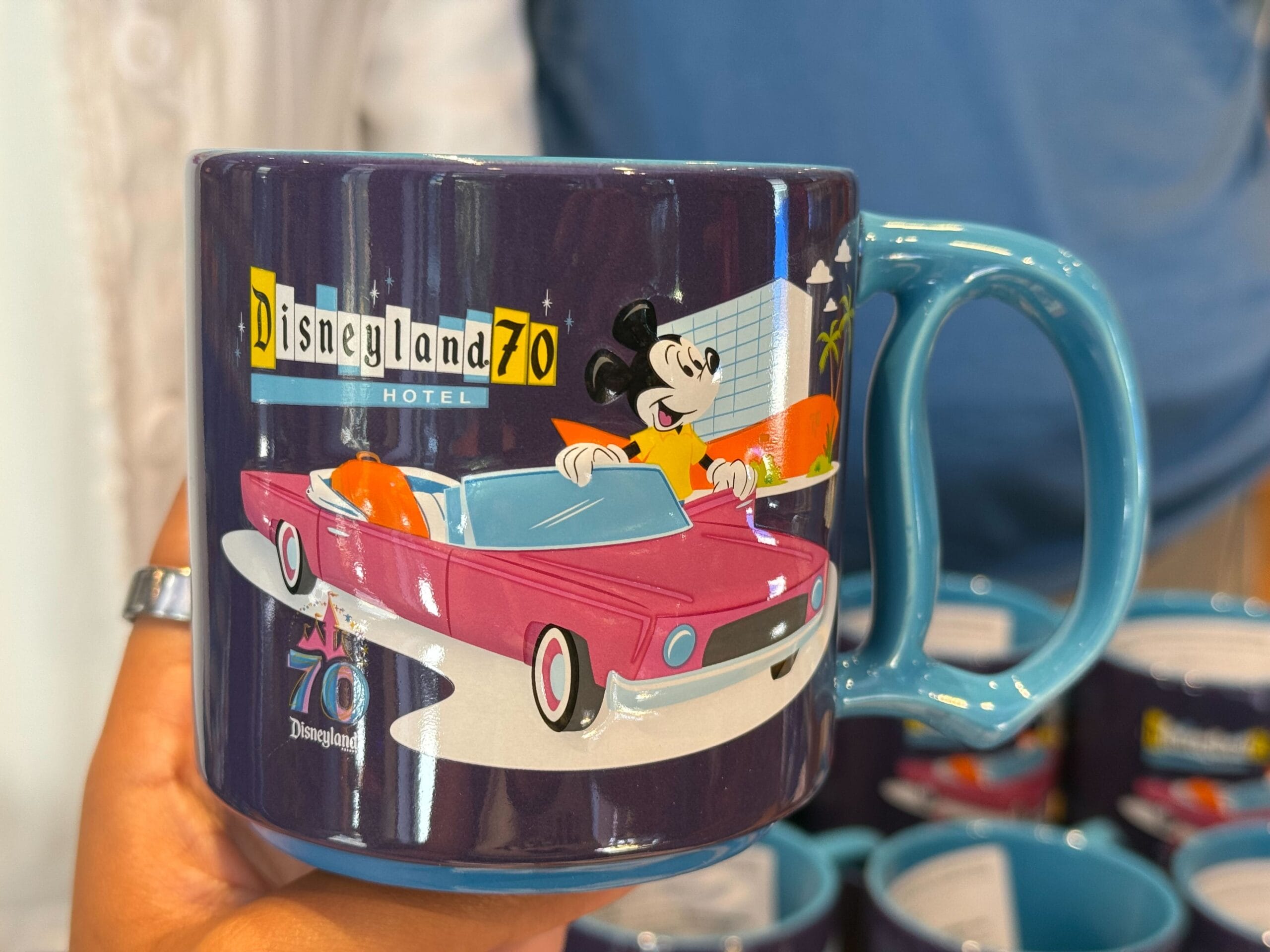 A hand holds a Disneyland Hotel 70th Anniversary mug with Mickey Mouse driving a pink car and palm trees in the background.