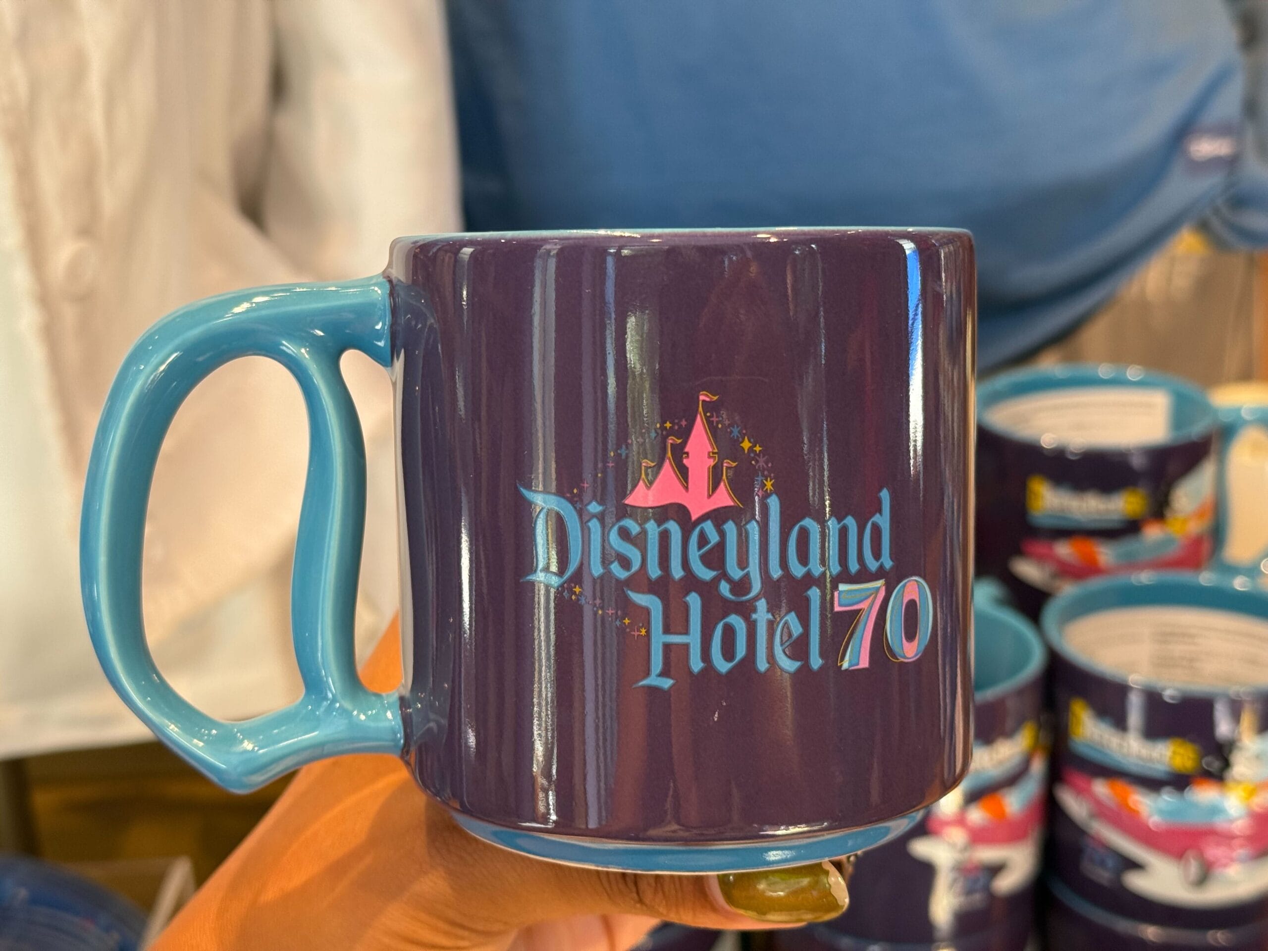 A hand holds a blue Disneyland Hotel 70th Anniversary mug from the Anniversary Collection with castle graphic and pastel lettering.