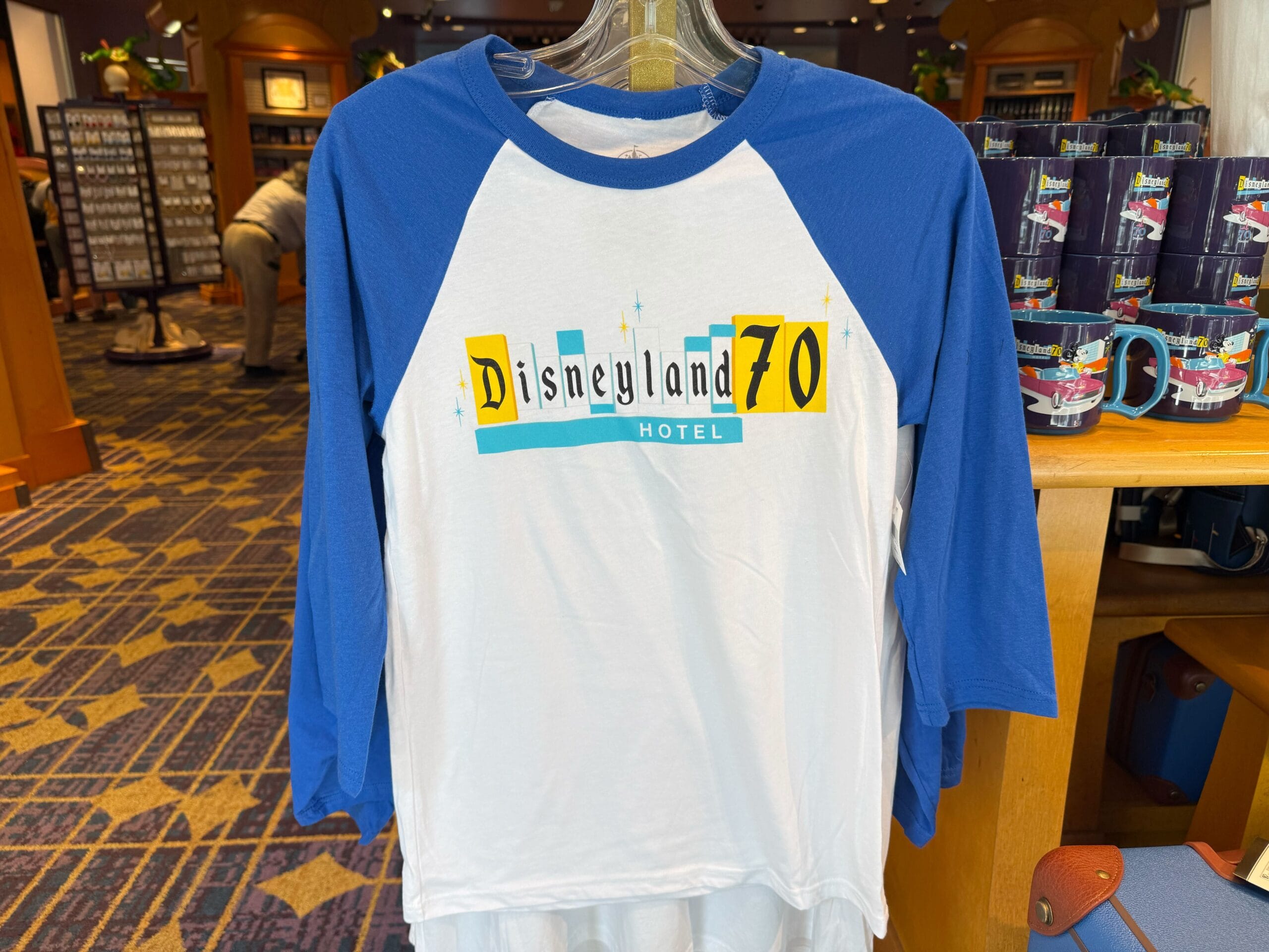 Disneyland Hotel 70th Anniversary Merchandise - White and blue Disneyland Hotel 70th Anniversary Collection shirt on a hanger in a gift shop.