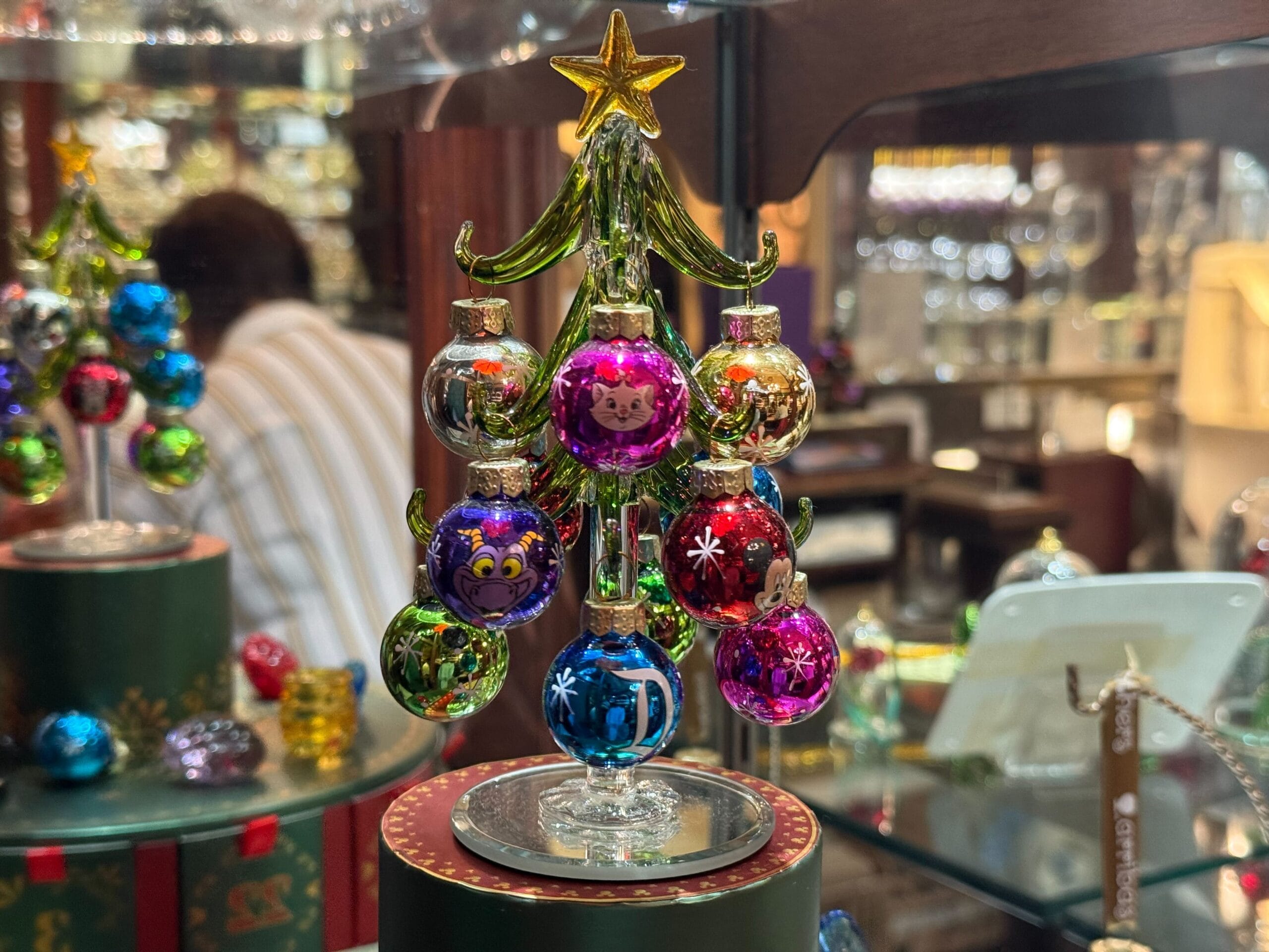 A small glass Christmas tree with colorful, Disney-style ornaments featuring cartoon faces, displayed indoors like in a Disney Park shop.