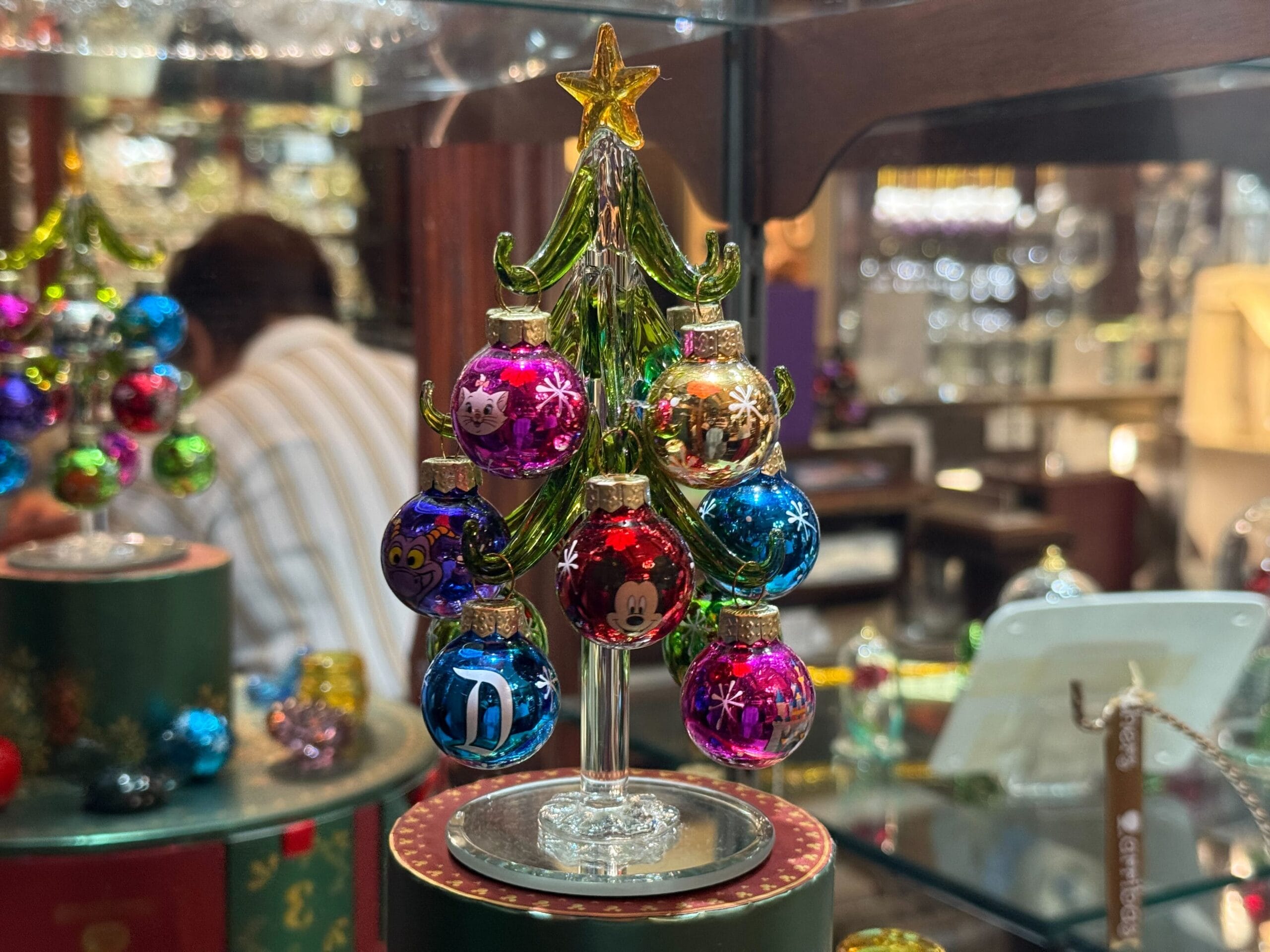 A small glass Christmas tree decorated with Disney-themed ornaments is showcased in a shop, evoking Disney Park holiday magic.