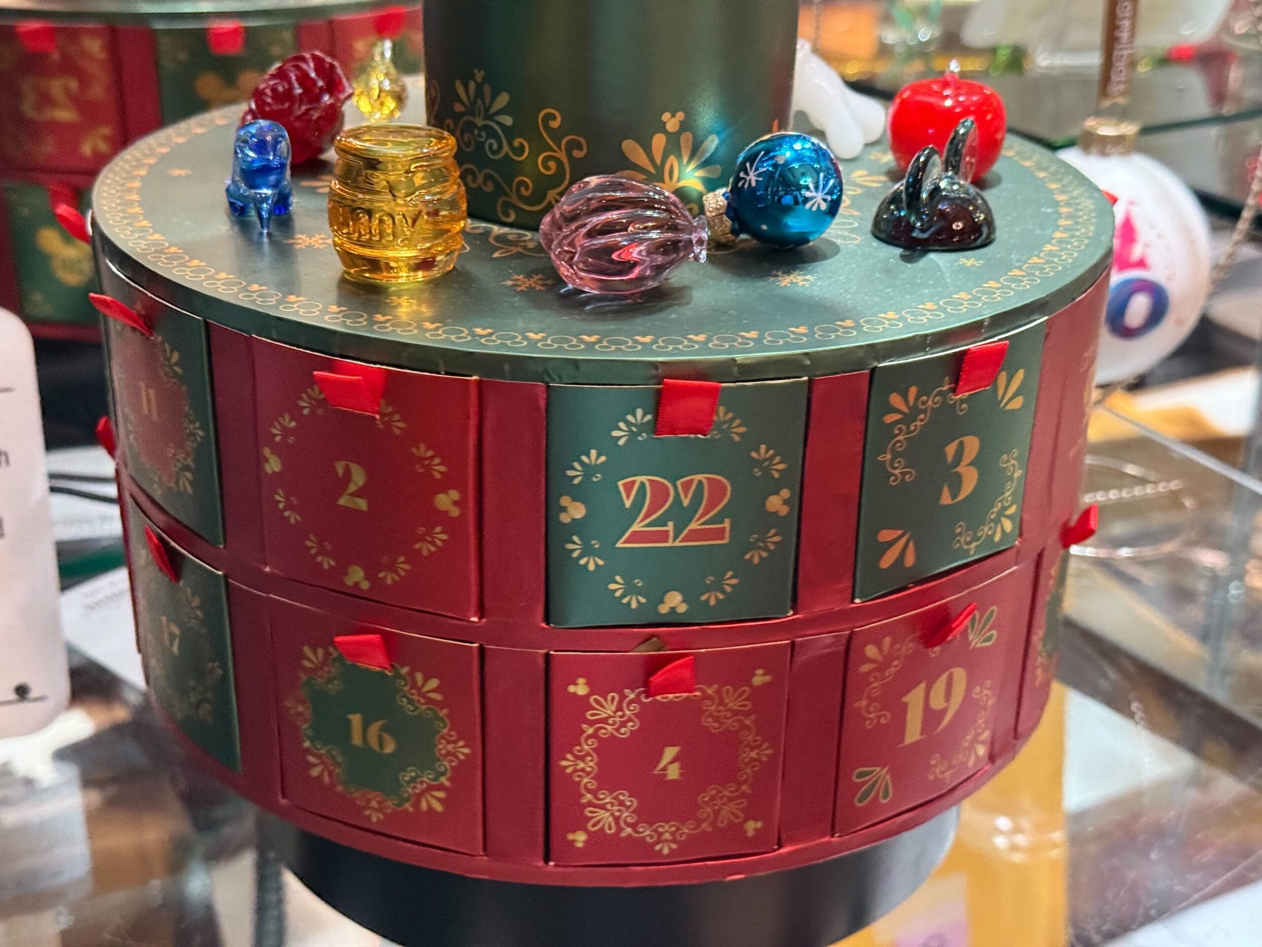 Round advent calendar with numbered drawers, topped with small colorful glass ornaments like Disney park holiday displays.