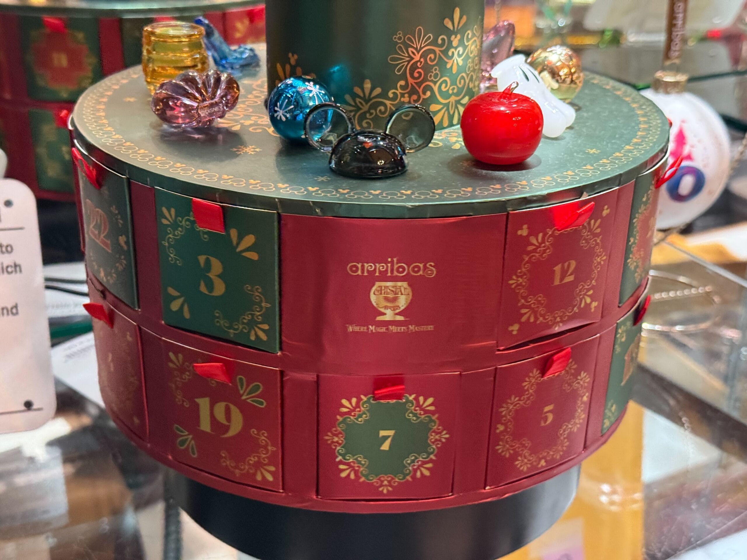 Festive round advent calendar with numbered red-green drawers, topped with glass ornaments and gold details, reminiscent of Disney holiday decor.