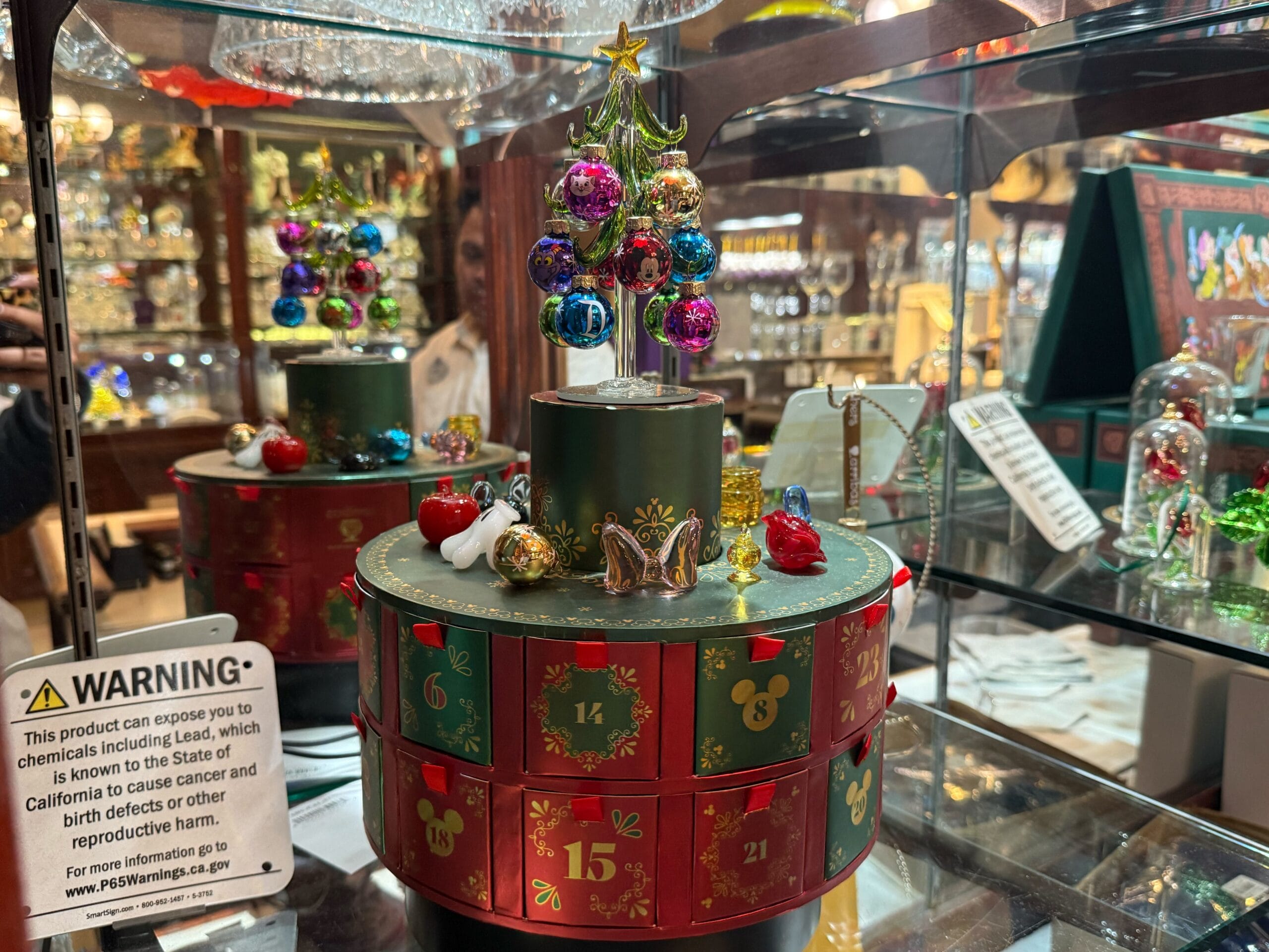 A festive Christmas advent calendar with a small tree and ornaments on top, displayed in a glass case like in a Disney park shop window.