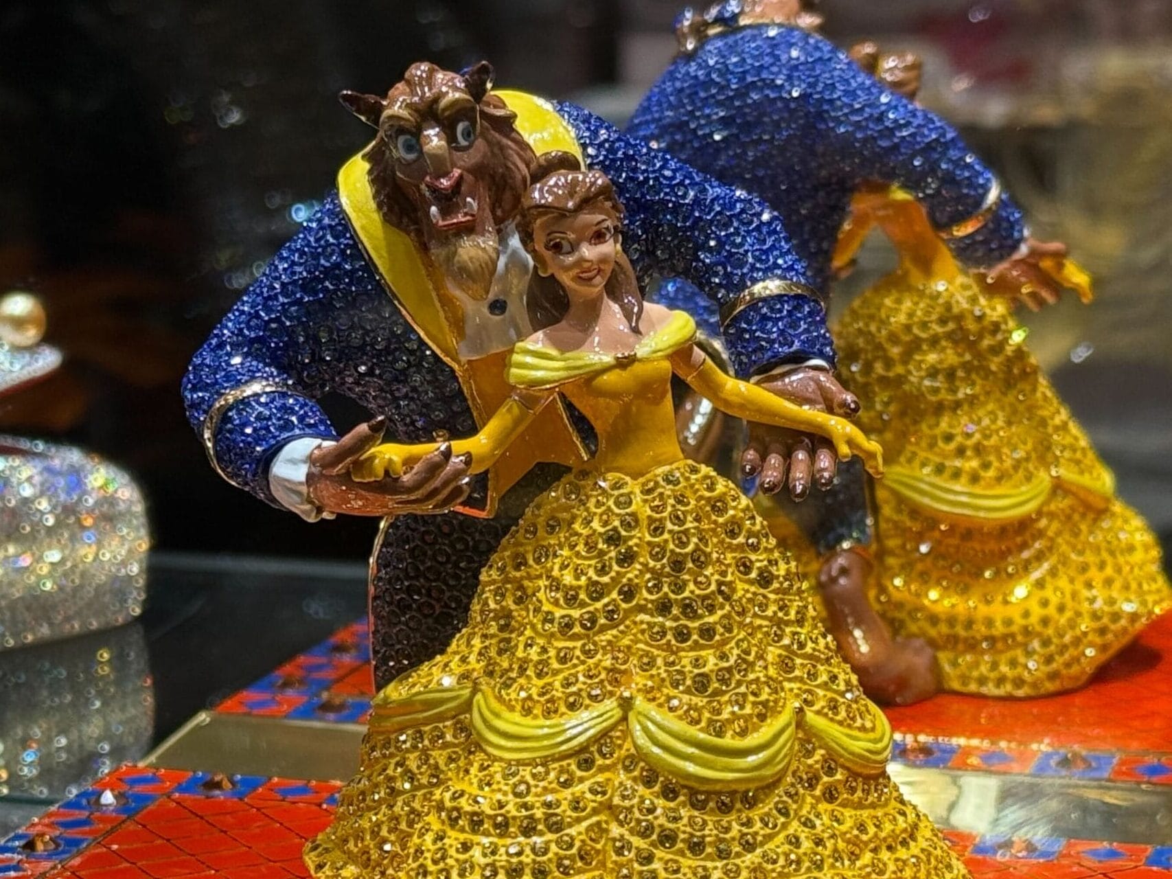 A jeweled Beauty and the Beast figurine on a red-blue tiled base, beside a glittery glass slipper, reminiscent of Disney Park displays.