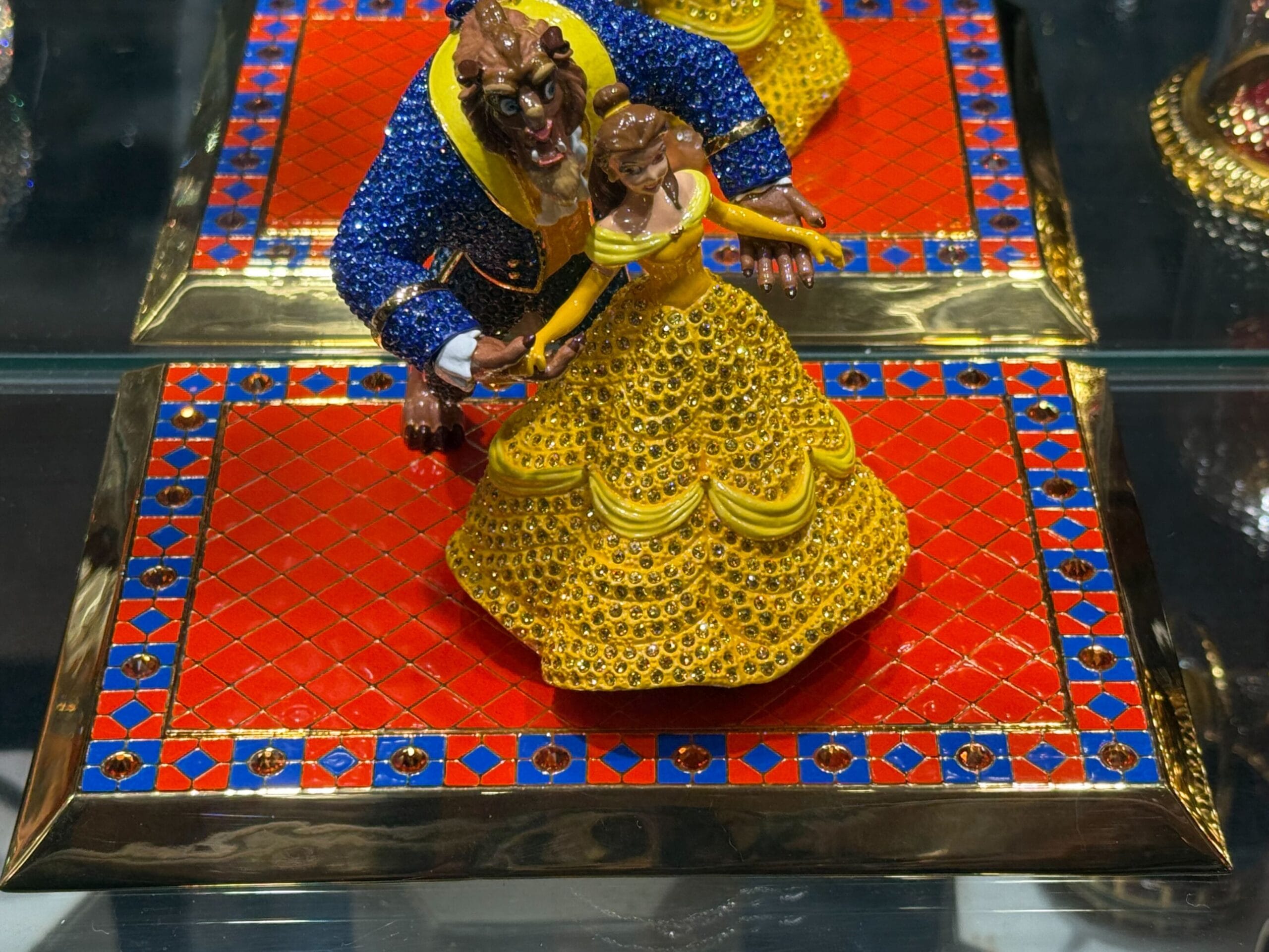 A jeweled Belle and Beast figurine dances on a red and gold tray, reminiscent of Disney Park merchandise from castle shops worldwide.
