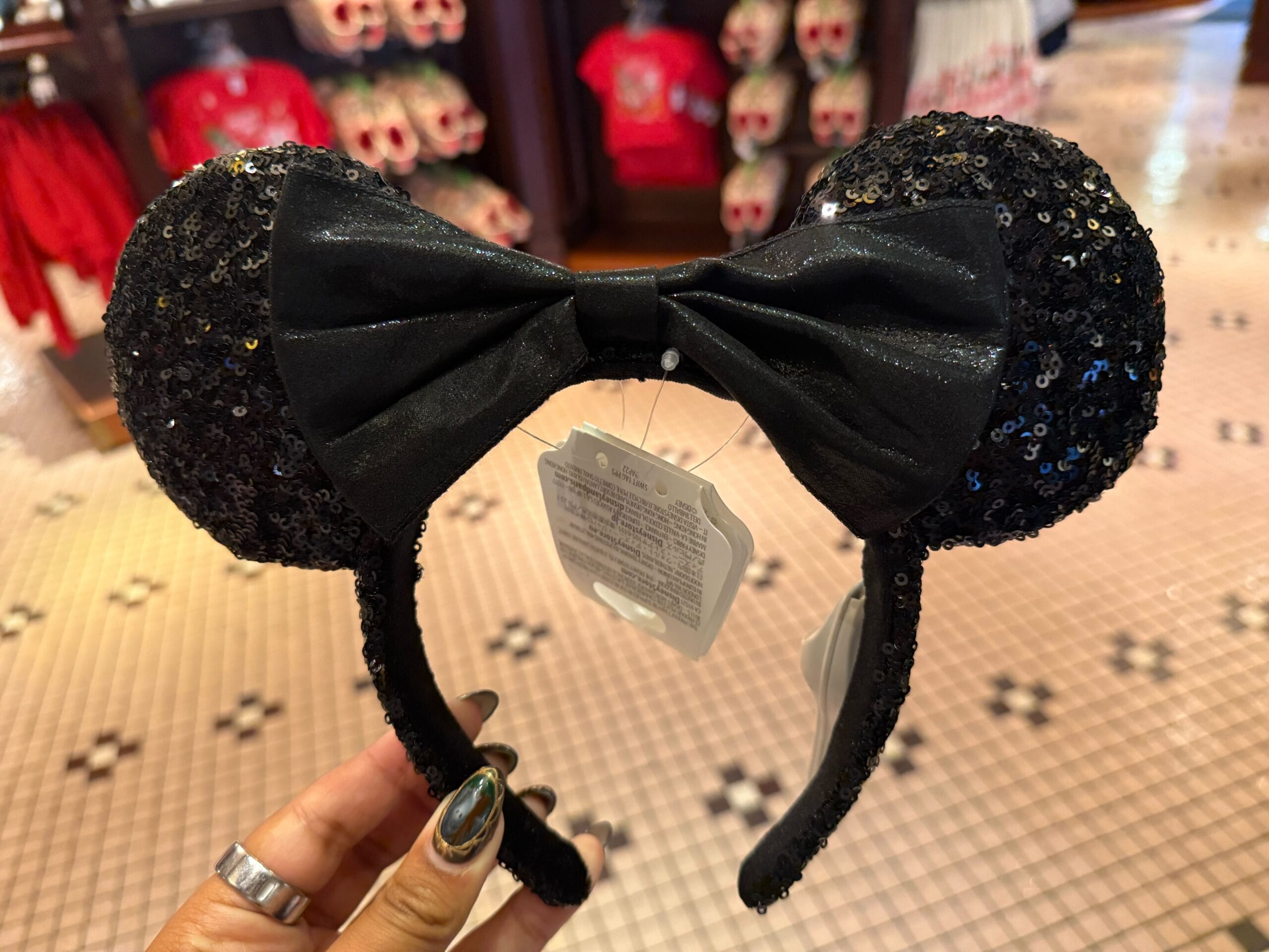 A hand holds a Black Sequined Minnie Ear Headband with a large black bow, displayed in a Disney World shop near Cinderella Castle.