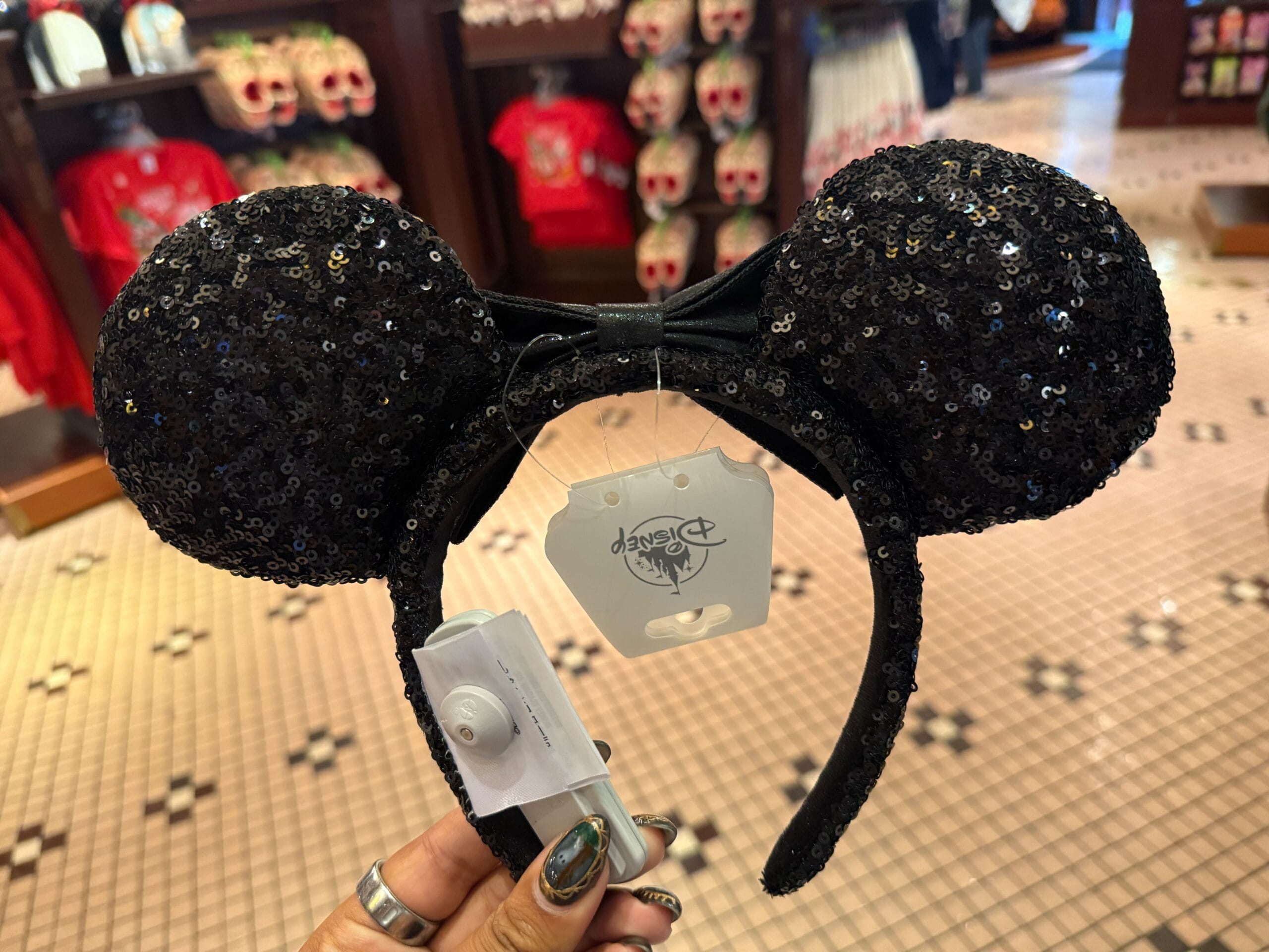 A hand displays a black sequined Minnie ear headband with a bow in a Disney World shop, pink floor tiles beneath, no castle in view.