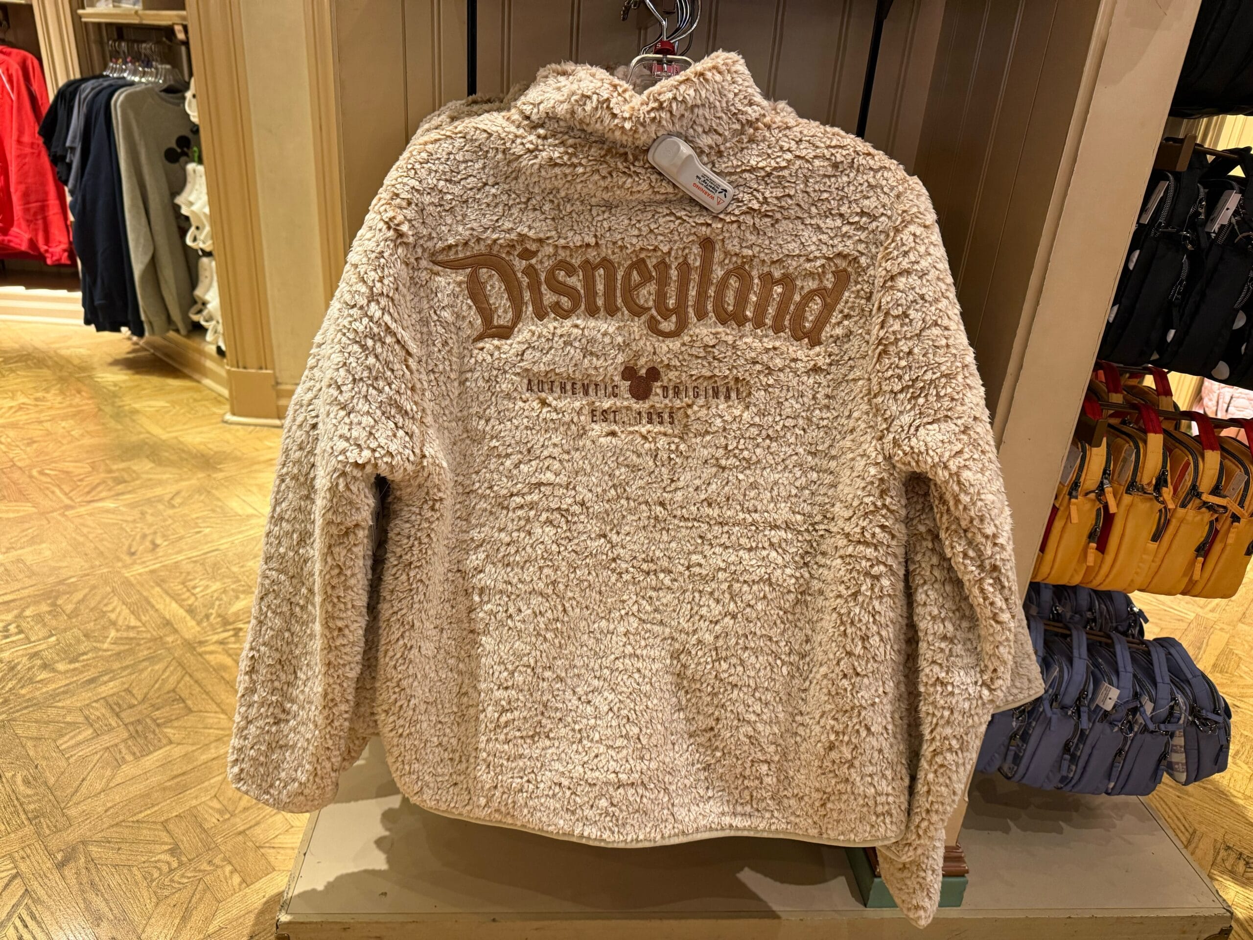 Cream-colored fuzzy Disneyland Logo Sweatshirt with brown lettering, hanging on a rack in a store.