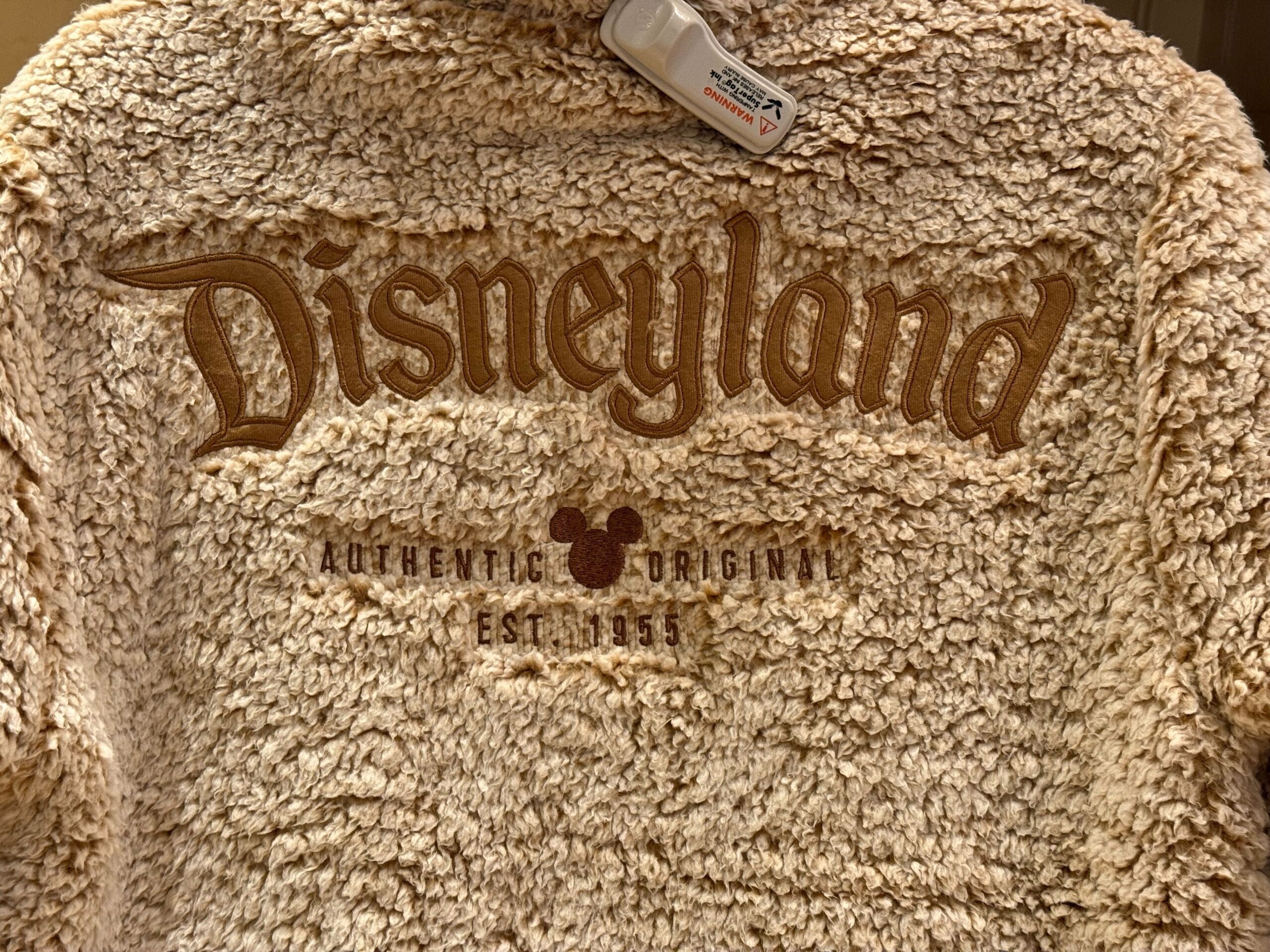 Beige fuzzy jacket with Disney logo, "Authentic Original Est. 1955," and Mickey icon—a cozy holiday Logo Sweatshirt.