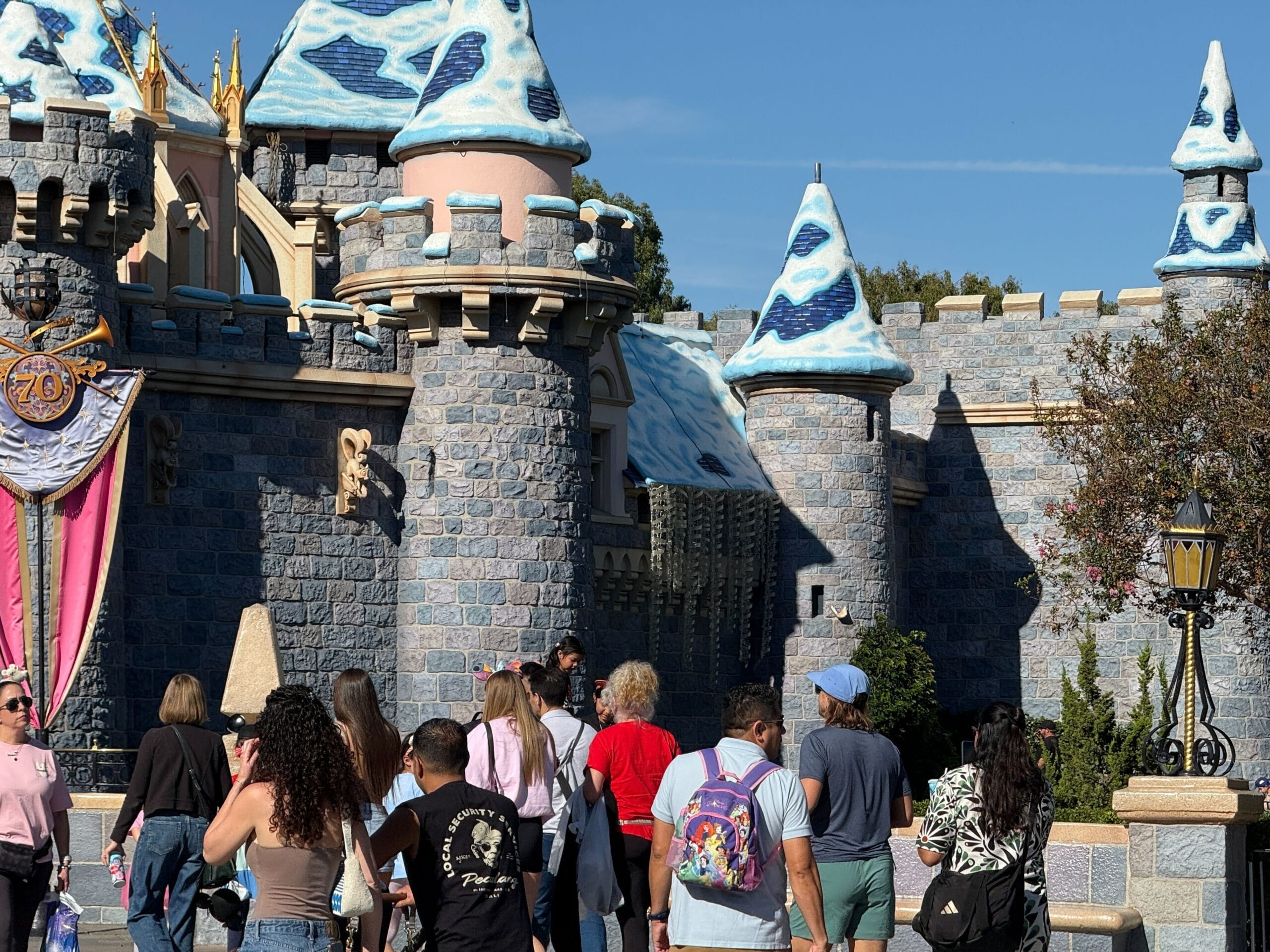 Guests approach a castle with blue and white turrets at Disneyland, resembling Sleeping Beauty Castle, under a bright sky.