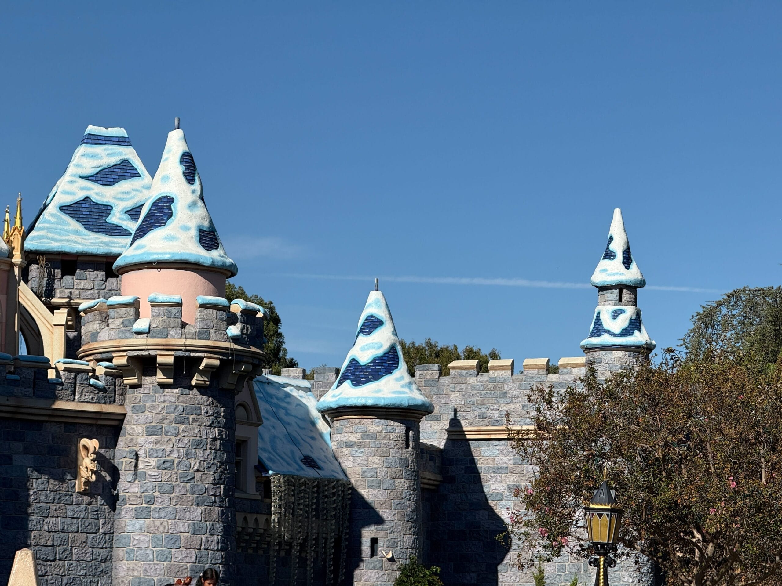 A castle with blue stone walls and snow-capped towers, resembling the iconic Disney castles, stands under a bright blue sky.