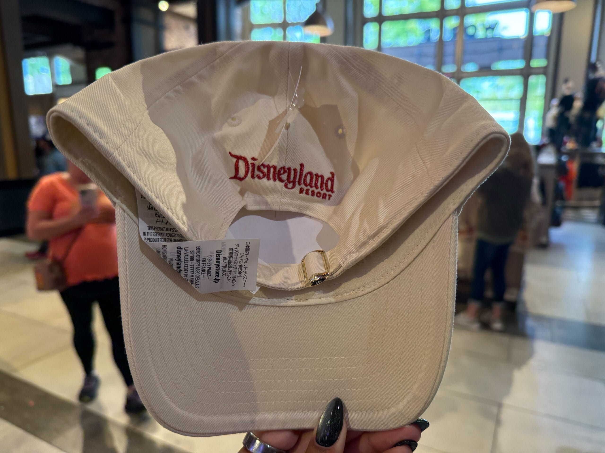 Inside a Disneyland store, someone holds up a beige Disneyland Resort cap, underside and tags visible—no castle in view.