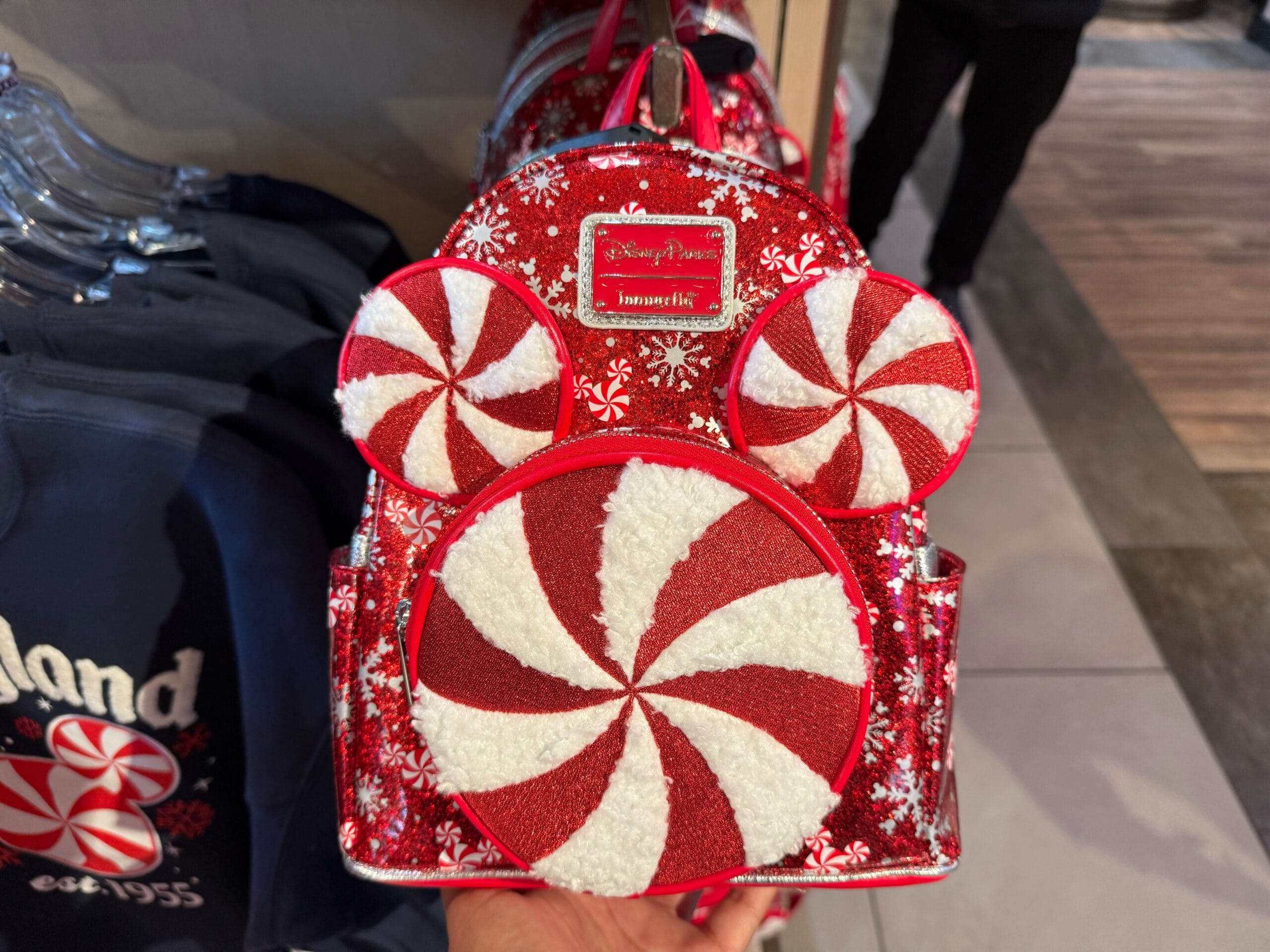 A red and white backpack with peppermint swirl designs on display in a Disney park shop, matching the festive park atmosphere.