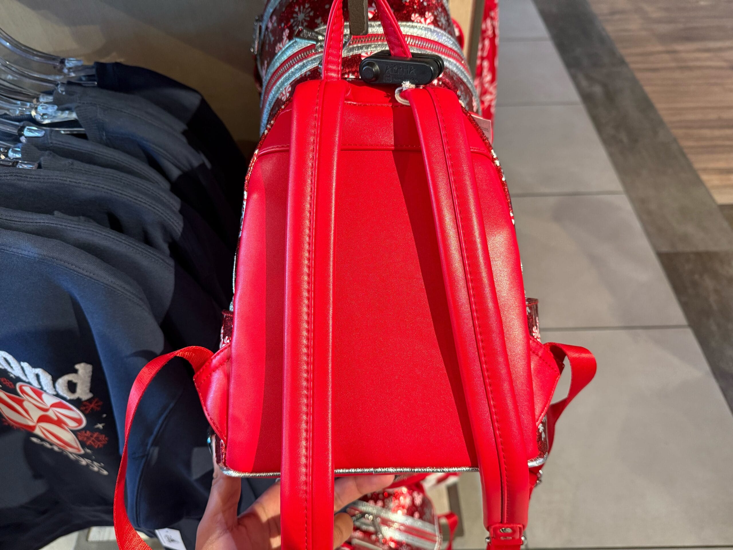 A bright red backpack with shiny trim sits on a store rack beside navy blue shirts, likely in a Disney park merchandise shop.