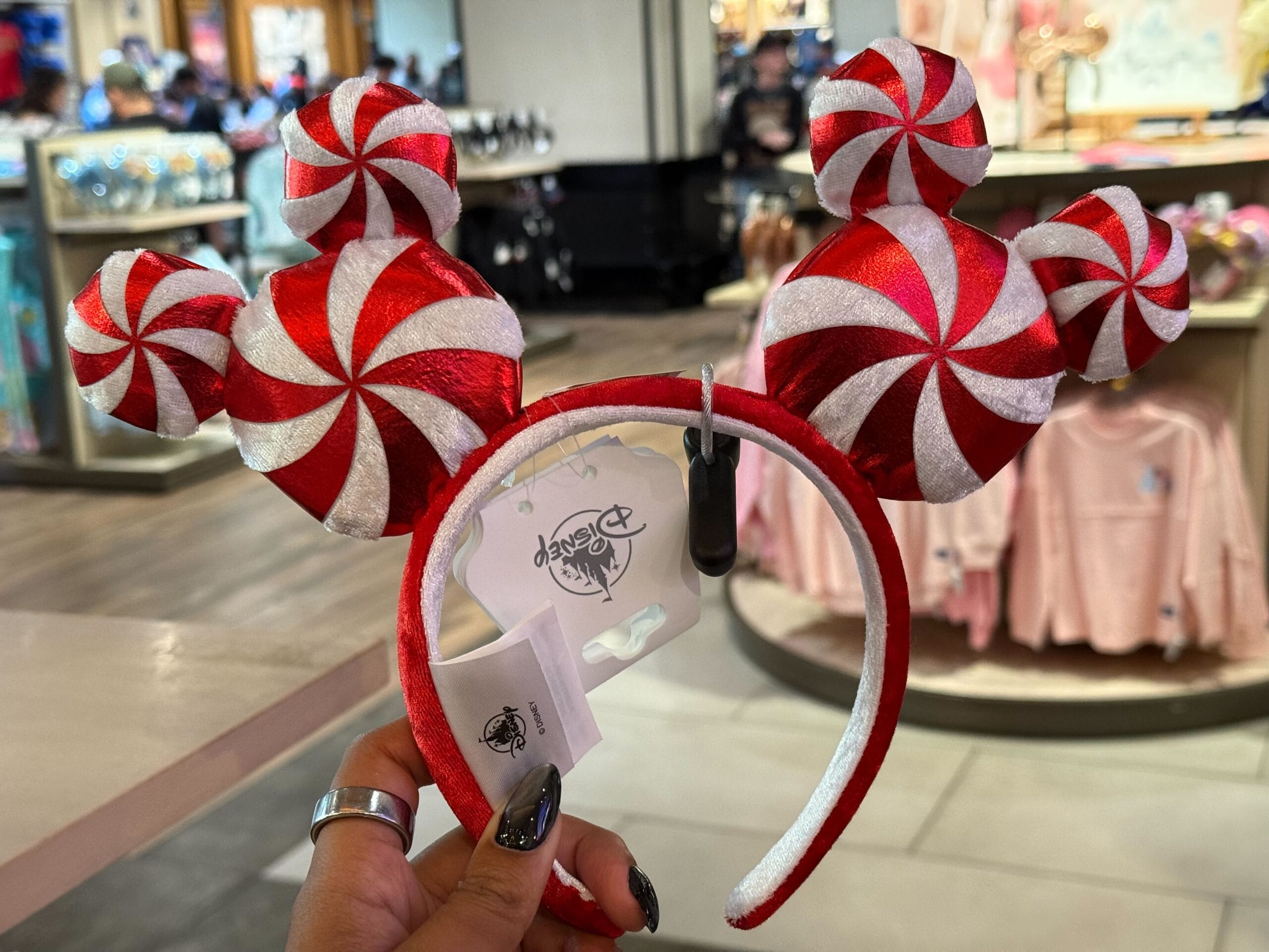 A hand holds a red and white peppermint Mickey Mouse ear headband inside a Disney park store, classic castle merch nearby.
