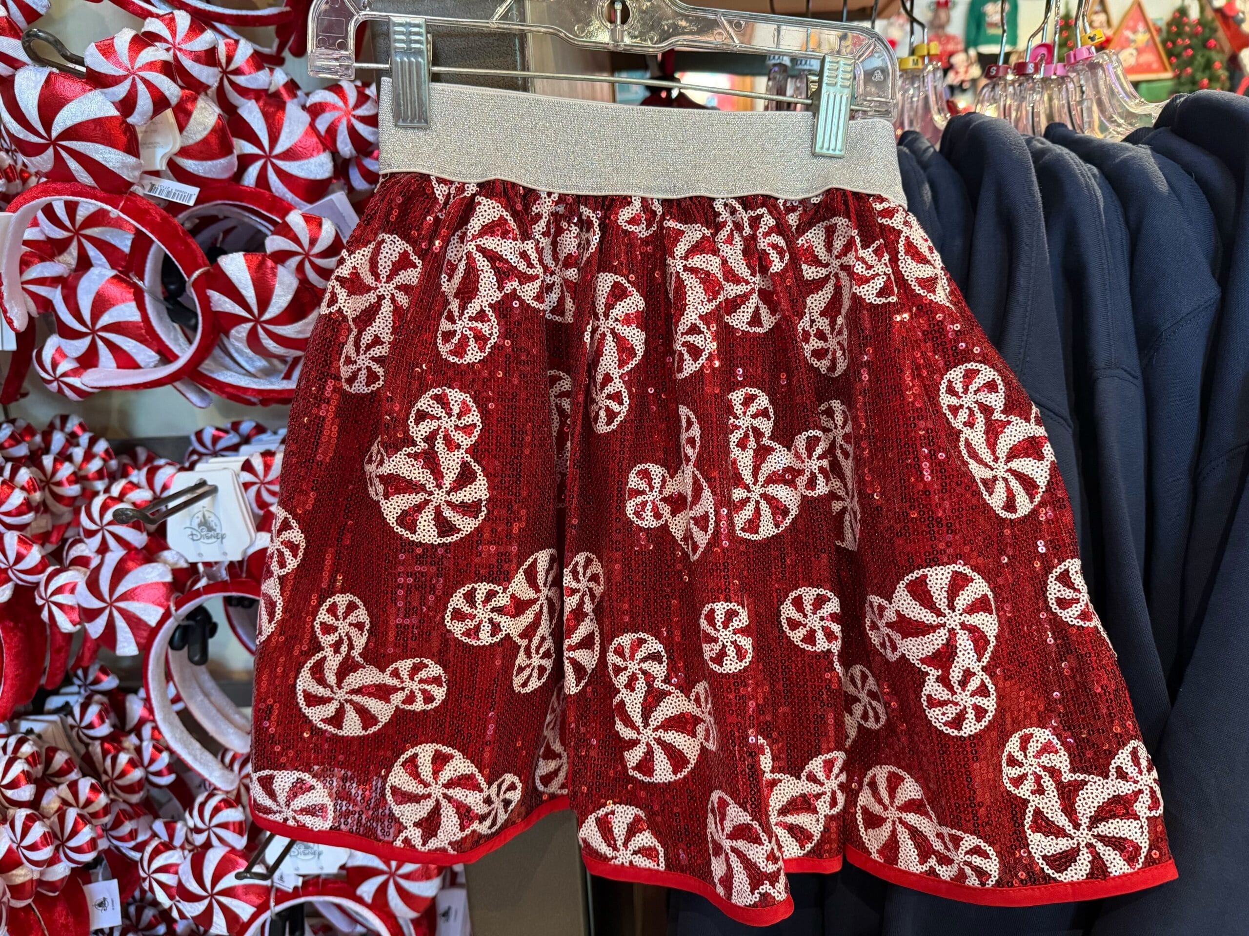Red sequin skirt with white peppermint candy patterns, likely in a Disney Park shop, hangs beside headbands and dark clothing.