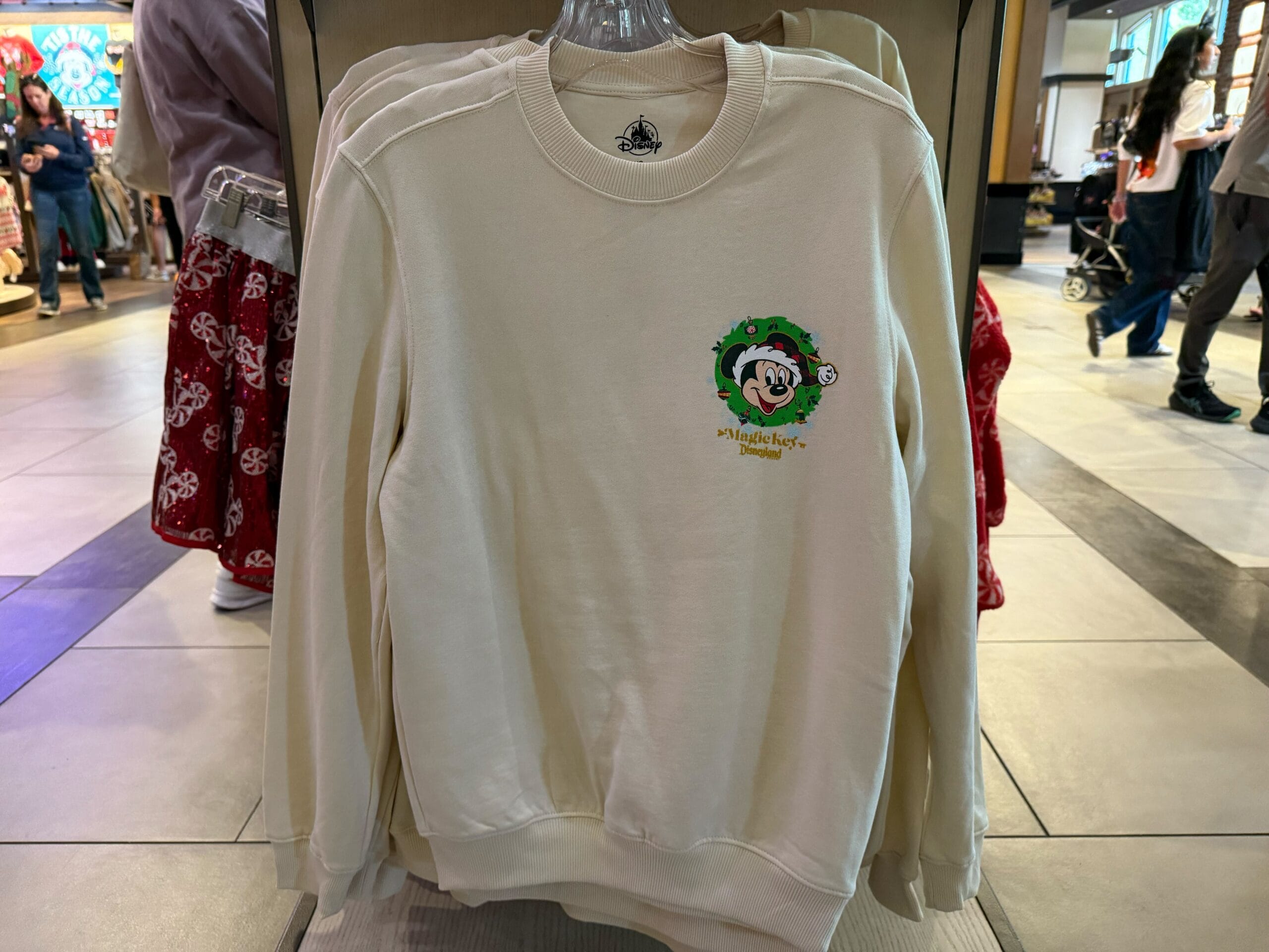 Cream Magic Key sweatshirt with Mickey Mouse in a holiday wreath, on a rack—merch you’d spot at Disneyland, not Disney World.