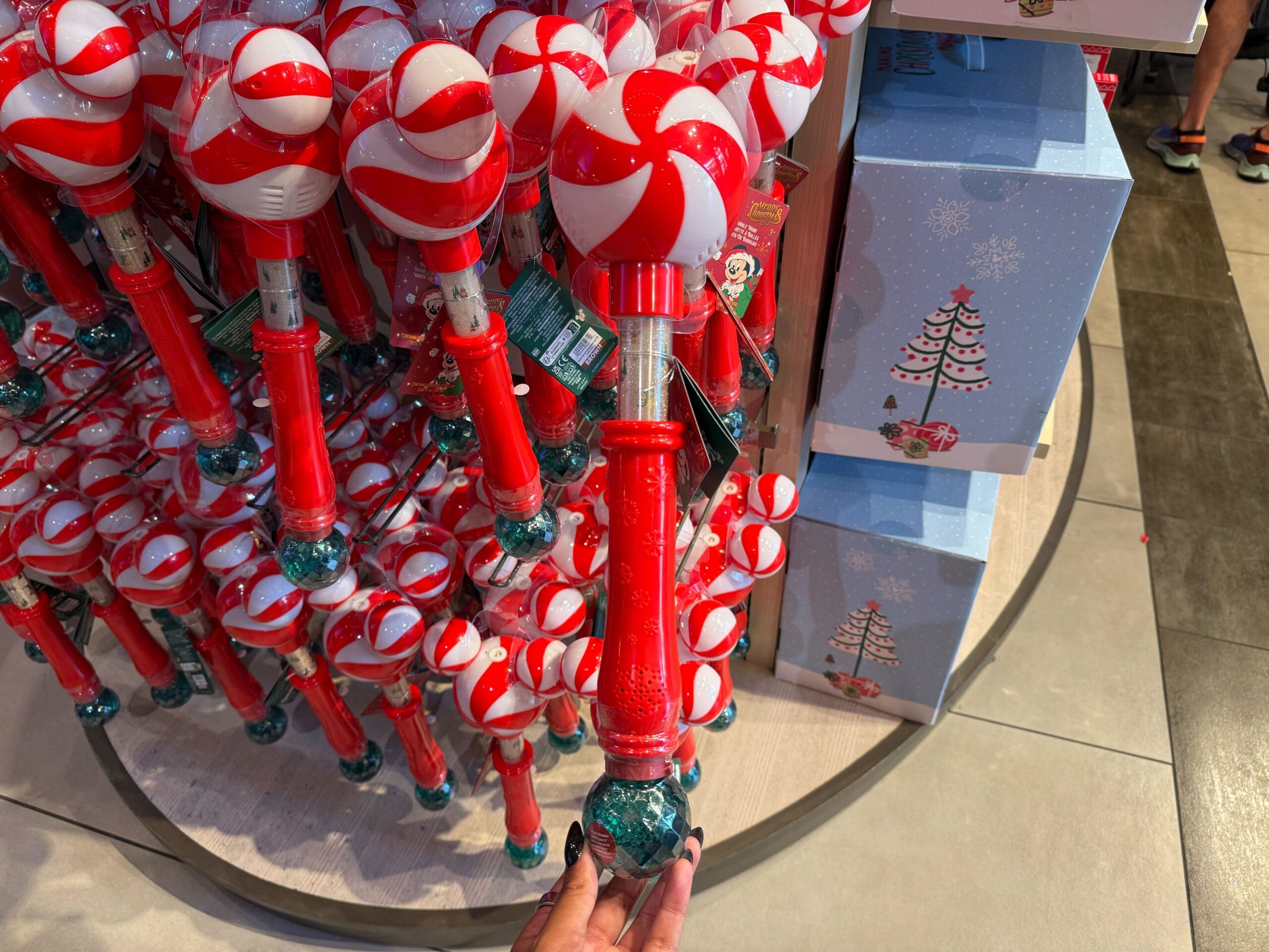 Red and white candy cane-themed toy wands on a shop shelf at a Disney park, hand holding a wand in the foreground.