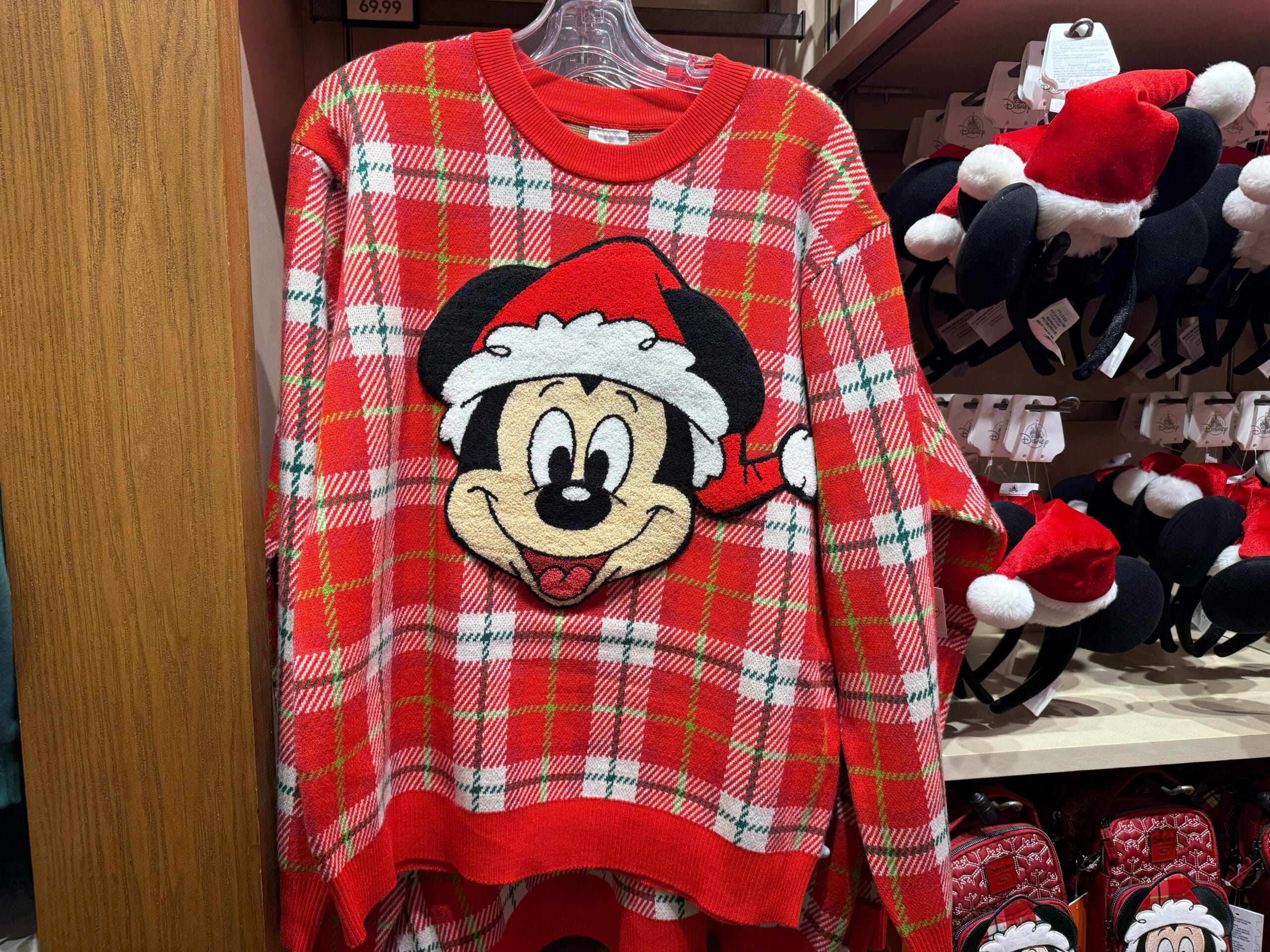 Red plaid Disneyland Christmas sweater with Mickey Mouse in a Santa hat, displayed in park merchandise store—no castle in view.