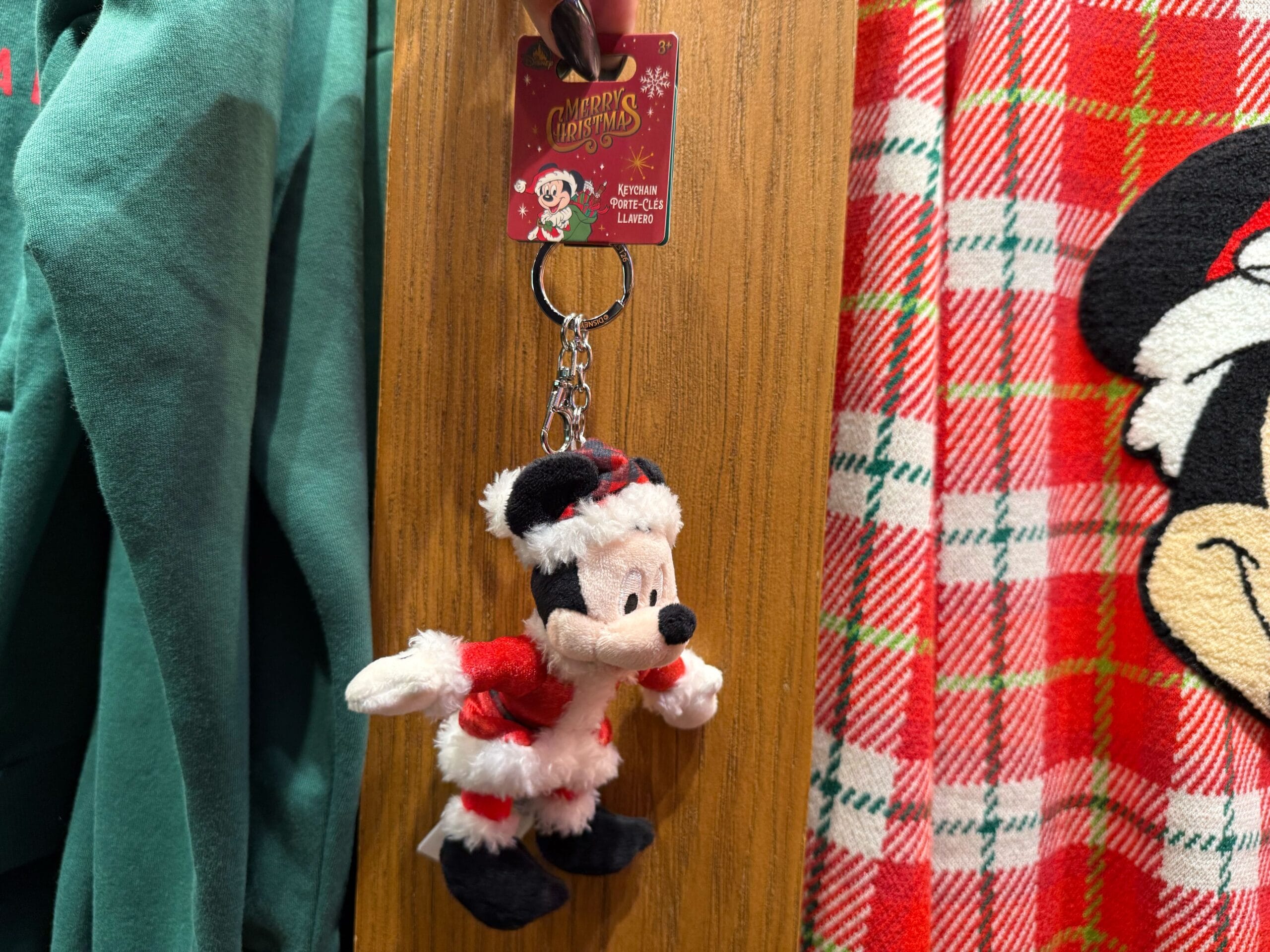 A plush Mickey Mouse Santa keychain hangs among Disneyland Christmas merchandise and festive outfits, Main Street castle not pictured.