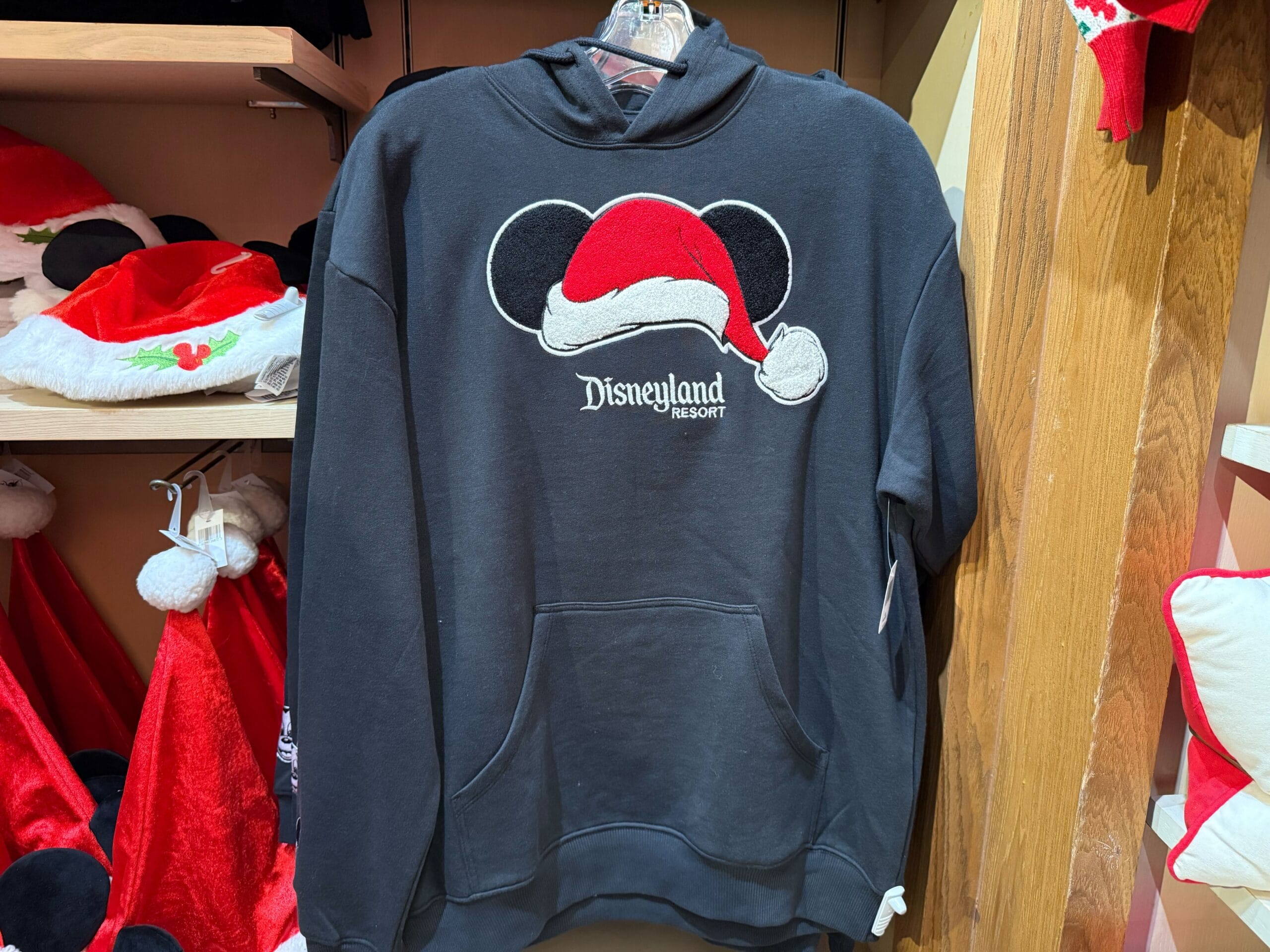Black Disneyland Resort hoodie with Mickey ears in a Santa hat, part of a holiday merch display at Disneyland, not Disney World.