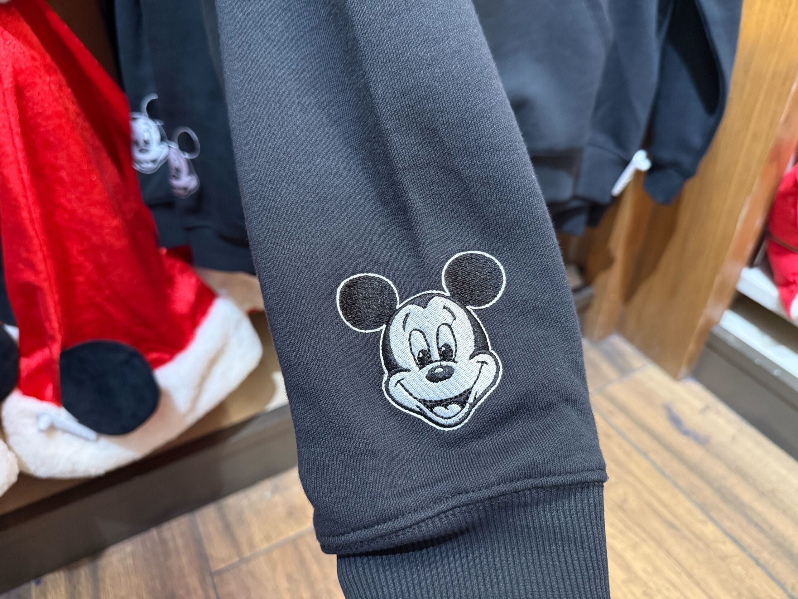 Black sleeve with embroidered Mickey Mouse face from the Peppermint Collection, surrounded by Disney Park apparel on wood floor.