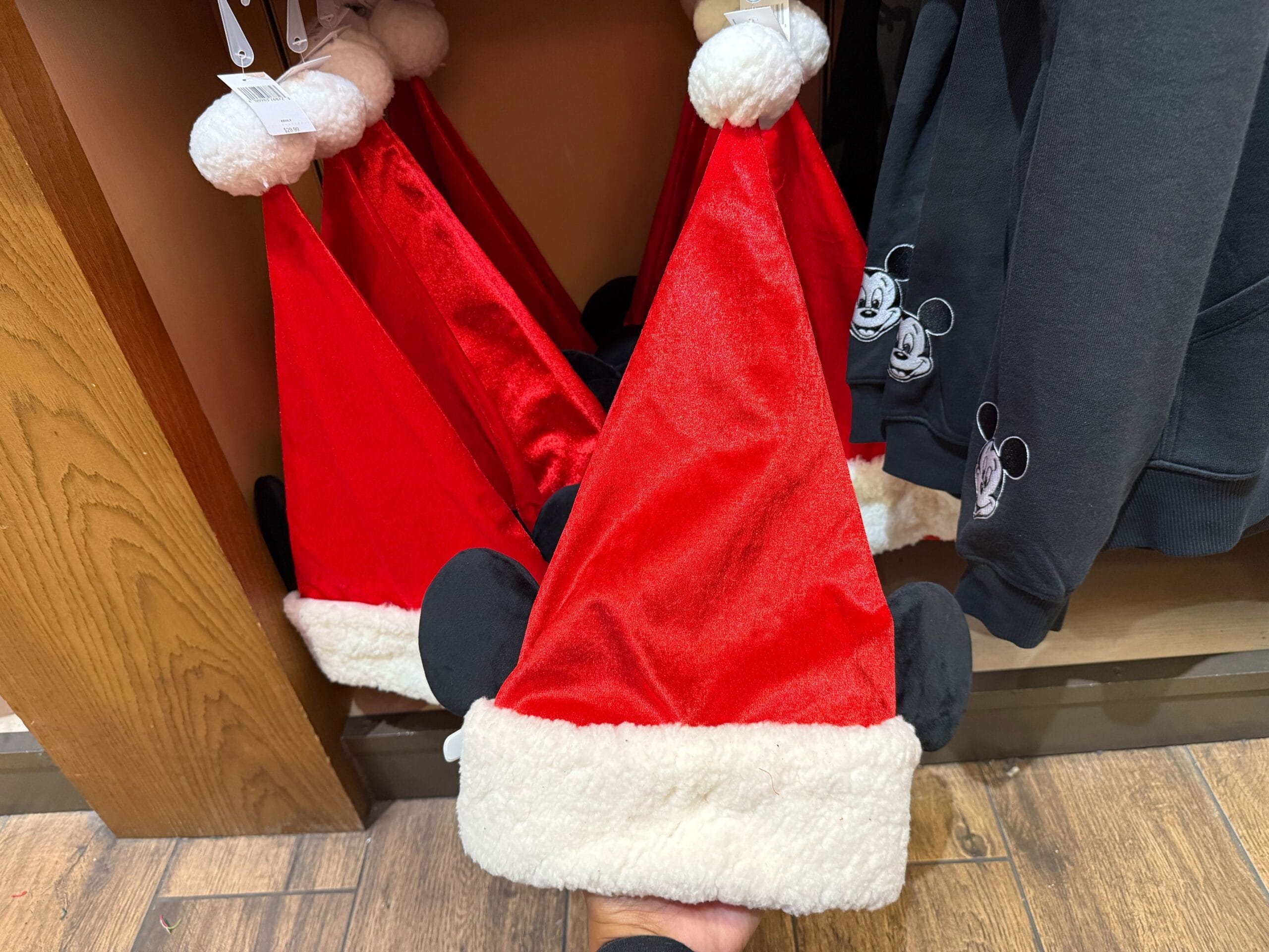 Red Santa hats with Mickey ears rest near a Magic Key sweatshirt in the Disneyland holiday display, in front of Sleeping Beauty Castle.