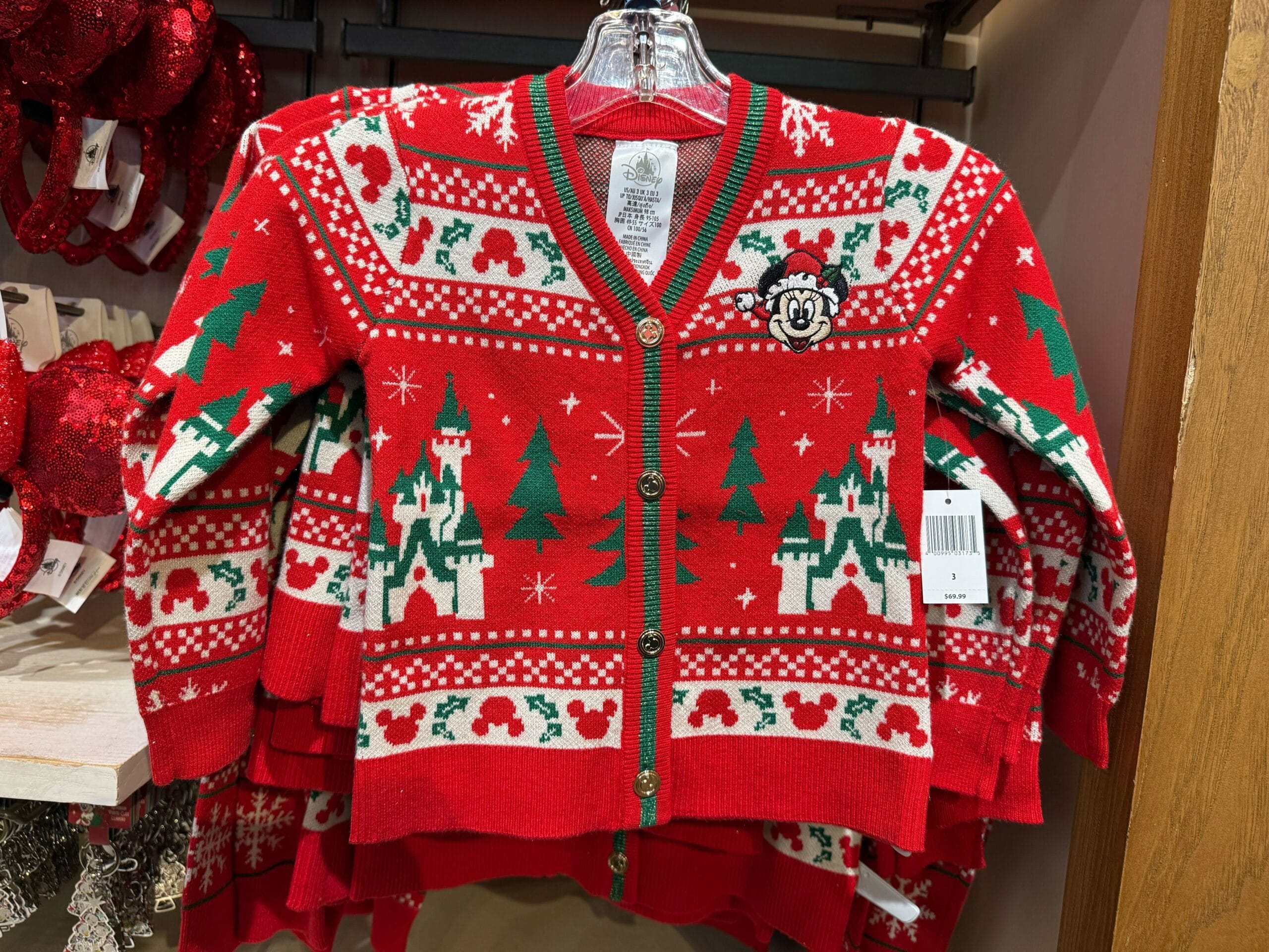 Red holiday sweater featuring green trees, snowflakes, and Disneyland castle resembling Sleeping Beauty Castle, plus Minnie Mouse patch.