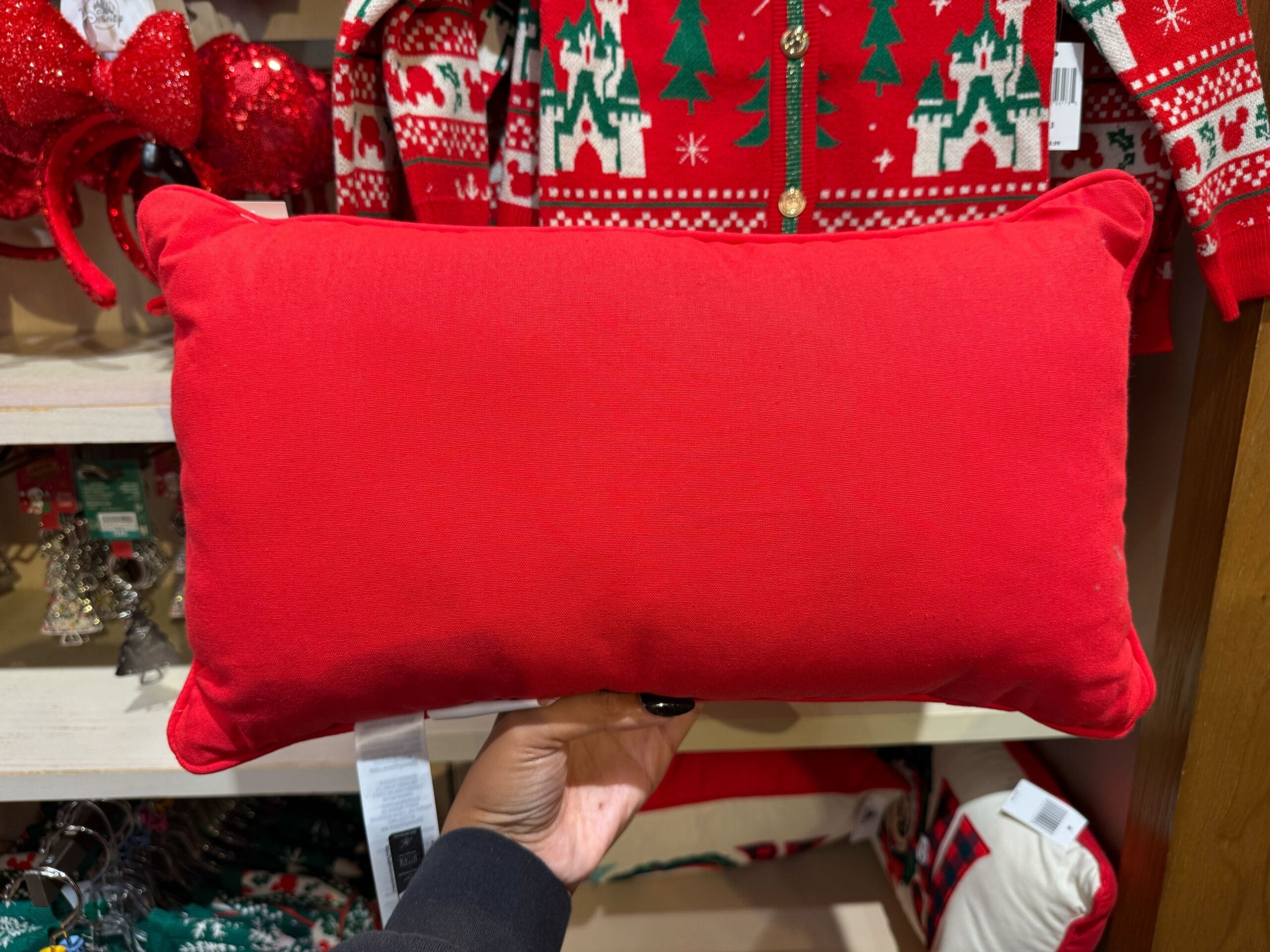 A hand holds a red pillow in a shop filled with Disneyland Christmas decor and festive sweaters, evoking park holiday magic.