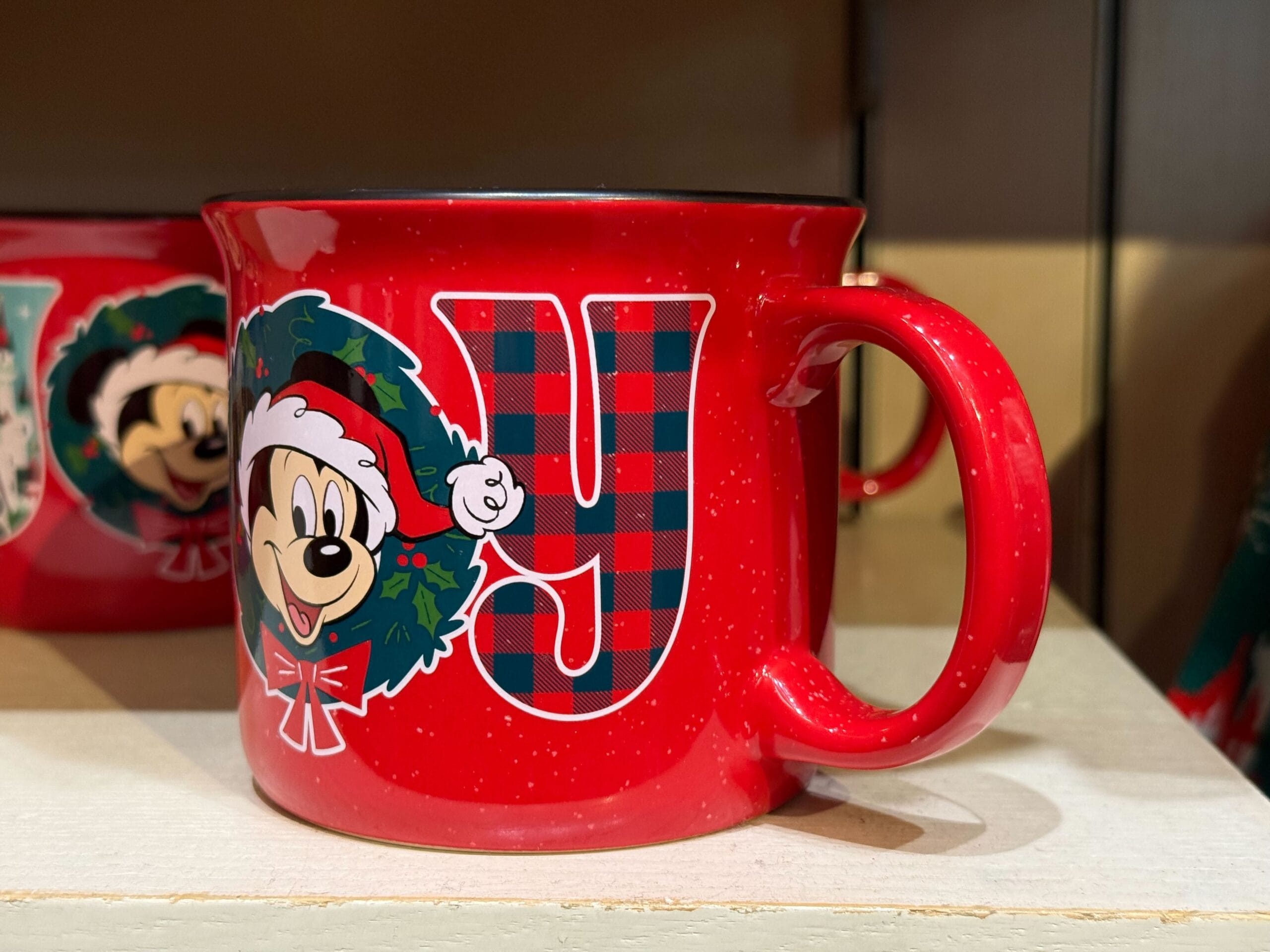 Red mug featuring Mickey Mouse in a holiday wreath with plaid "Y," part of Disneyland's Christmas collection on display.