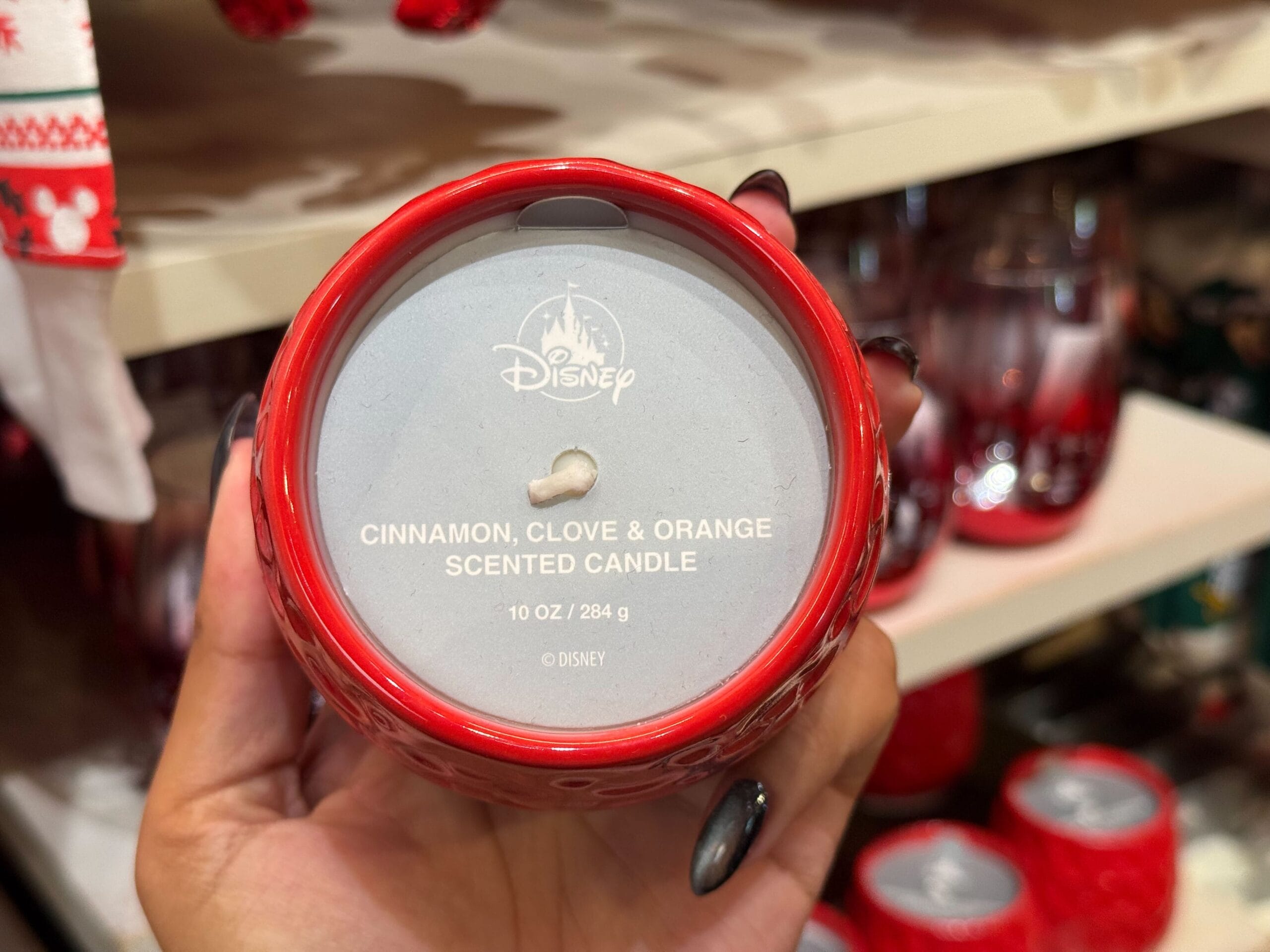 A hand holds a red Disney candle from Disneyland’s Christmas line, labeled "Cinnamon, Clove & Orange," no castle in view.