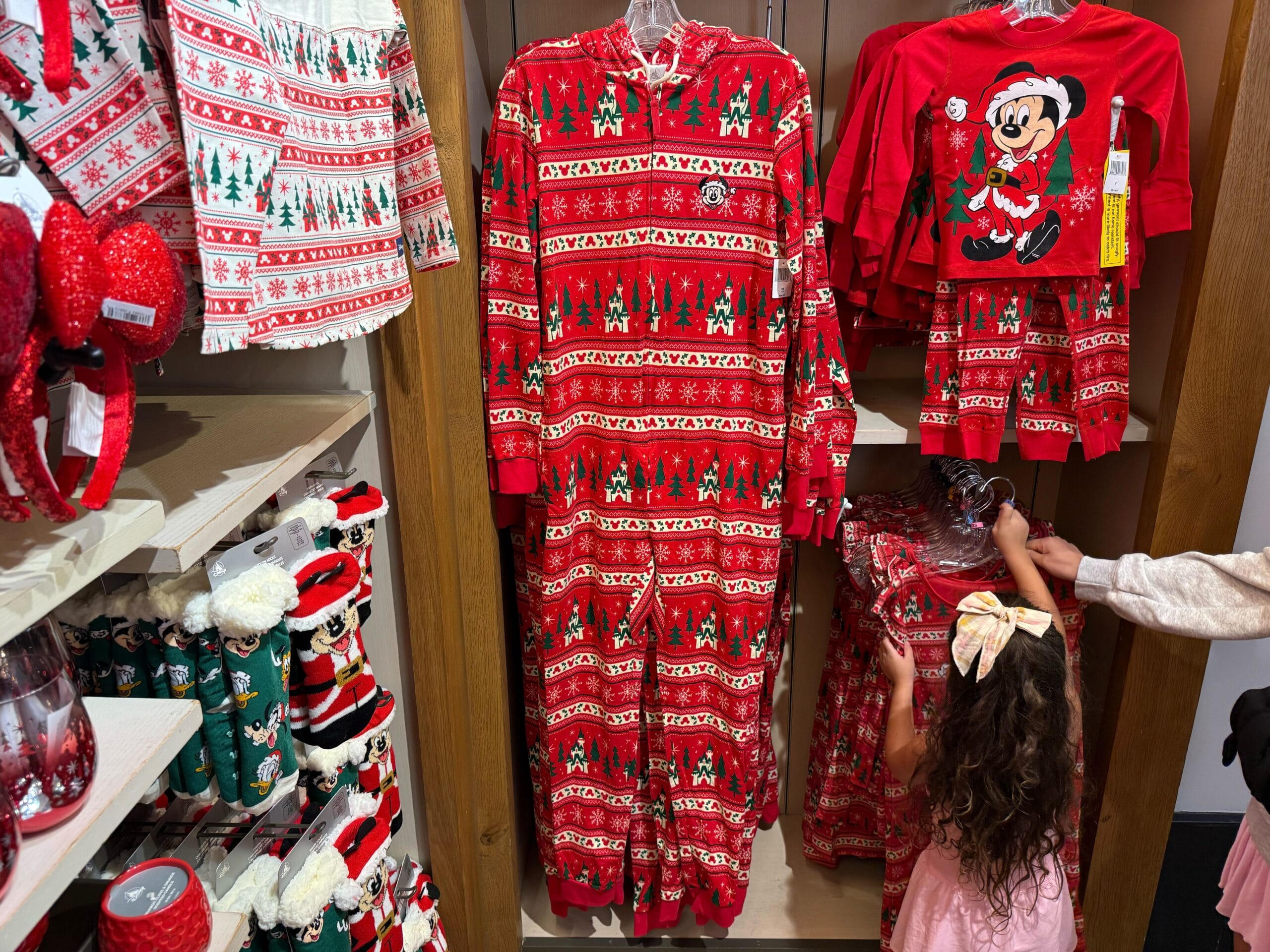 A child admires Disneyland Christmas merchandise, like red Mickey Mouse pajamas, on shelves—Sleeping Beauty Castle visible nearby.
