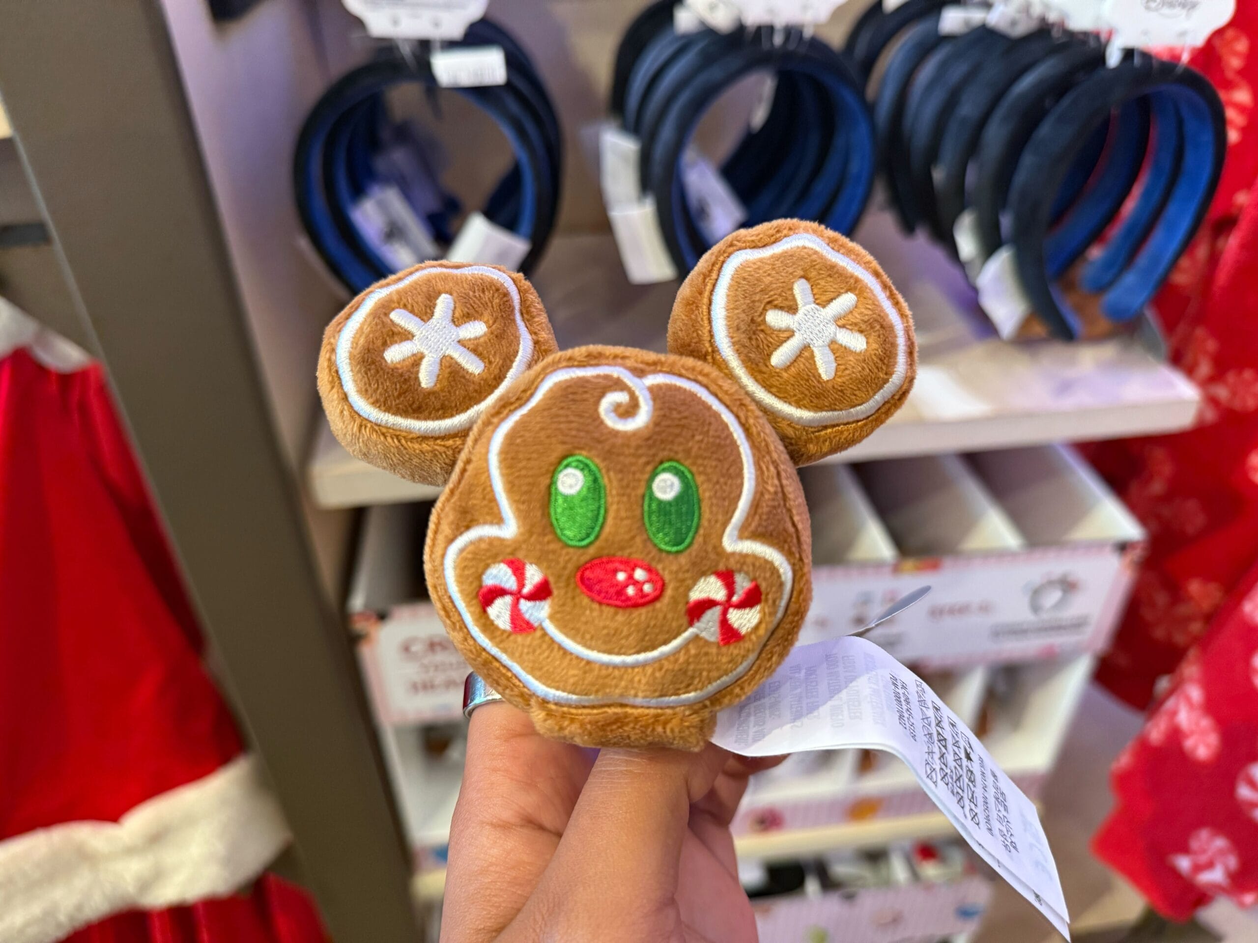 A hand holds a plush gingerbread Mickey Mouse headband from Disneyland’s holiday merch, surrounded by festive store displays.
