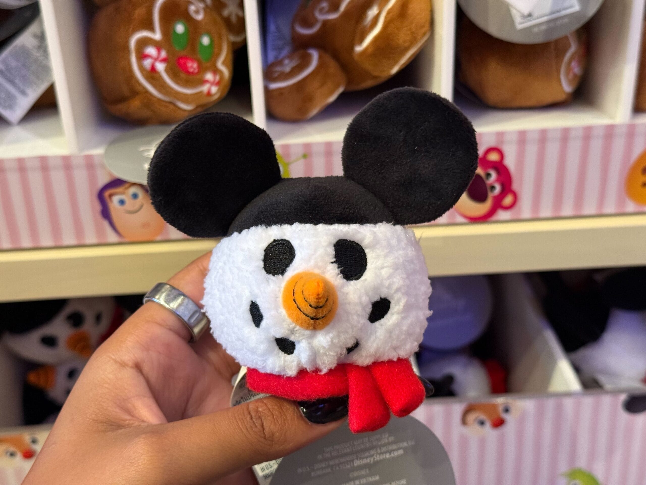 A hand holds a plush snowman with mouse ears from Disneyland Christmas merch, displayed in a festive store near Disney Park décor.