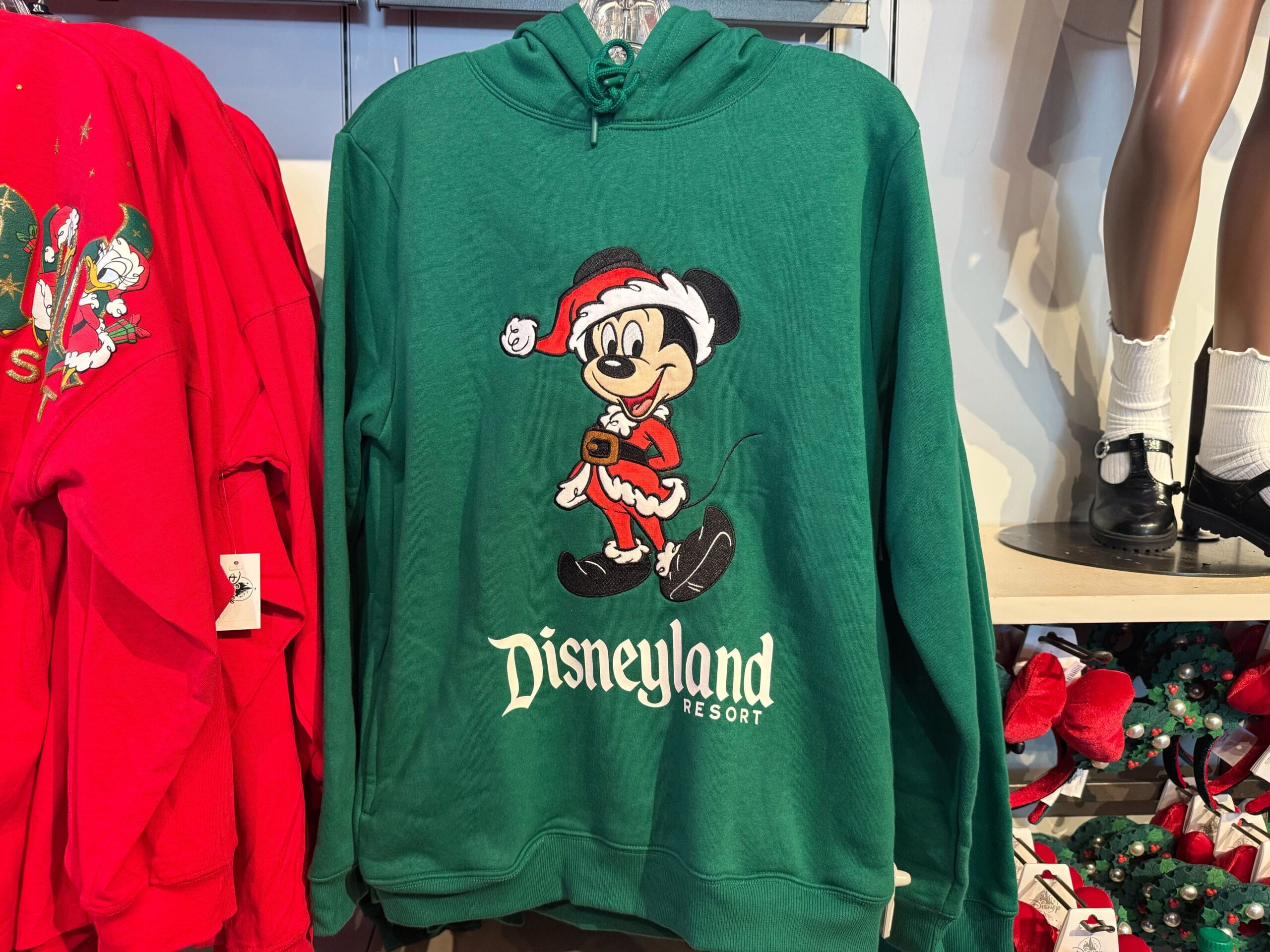 Green Disneyland holiday hoodie with Mickey as Santa, displayed in a Disney park shop—cozy festive merch, no castle visible.