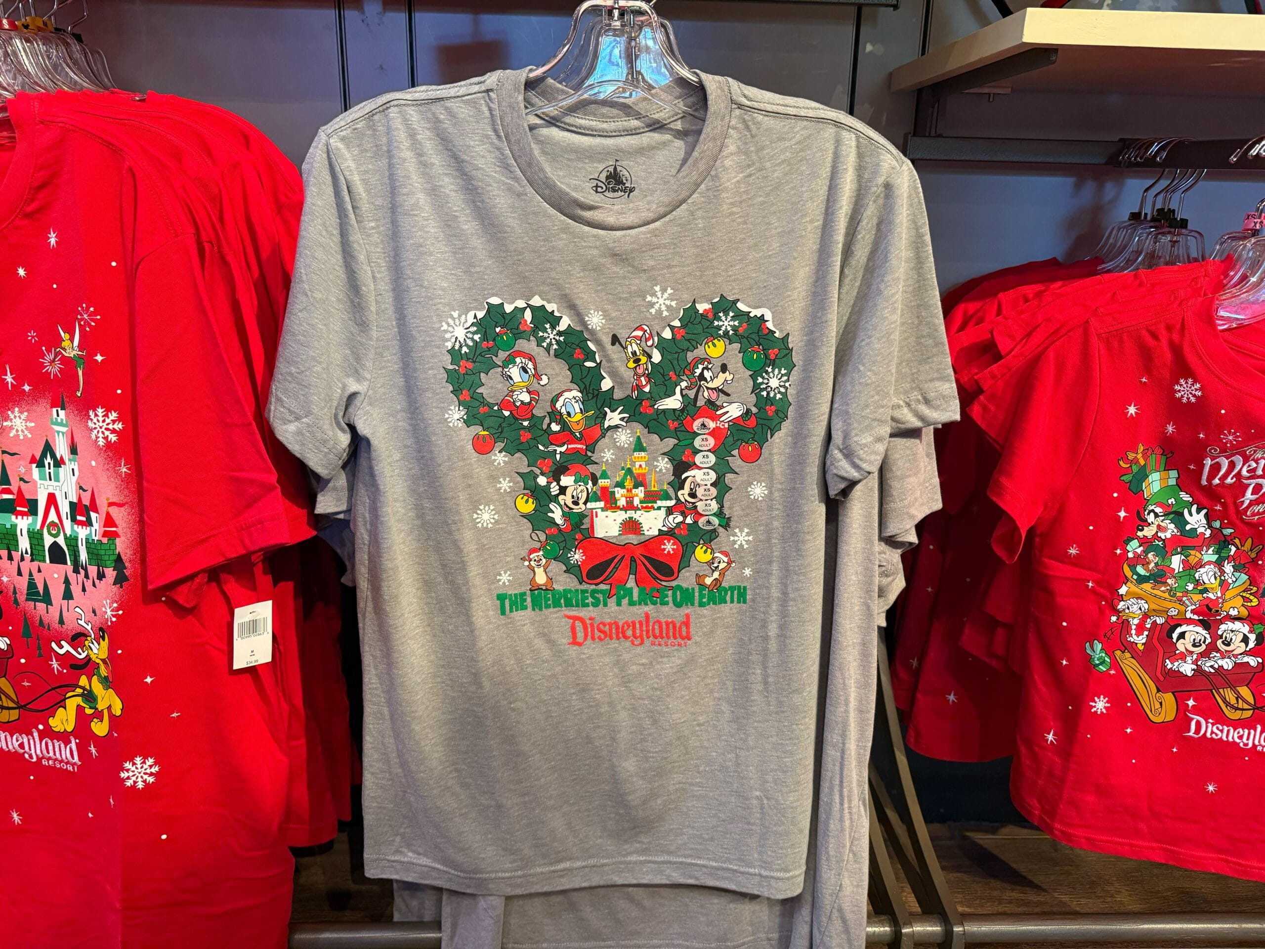 Gray Disneyland Christmas shirt with Mickey wreath, surrounded by festive red shirts, on display in the park’s holiday shop.