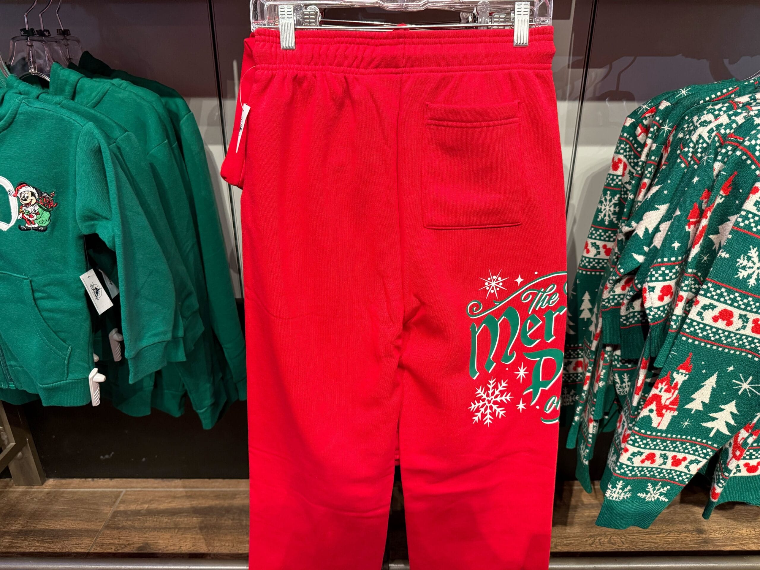 Bright red Disneyland Christmas sweatpants with "The Merry" and snowflakes, nestled among festive green park holiday sweaters.