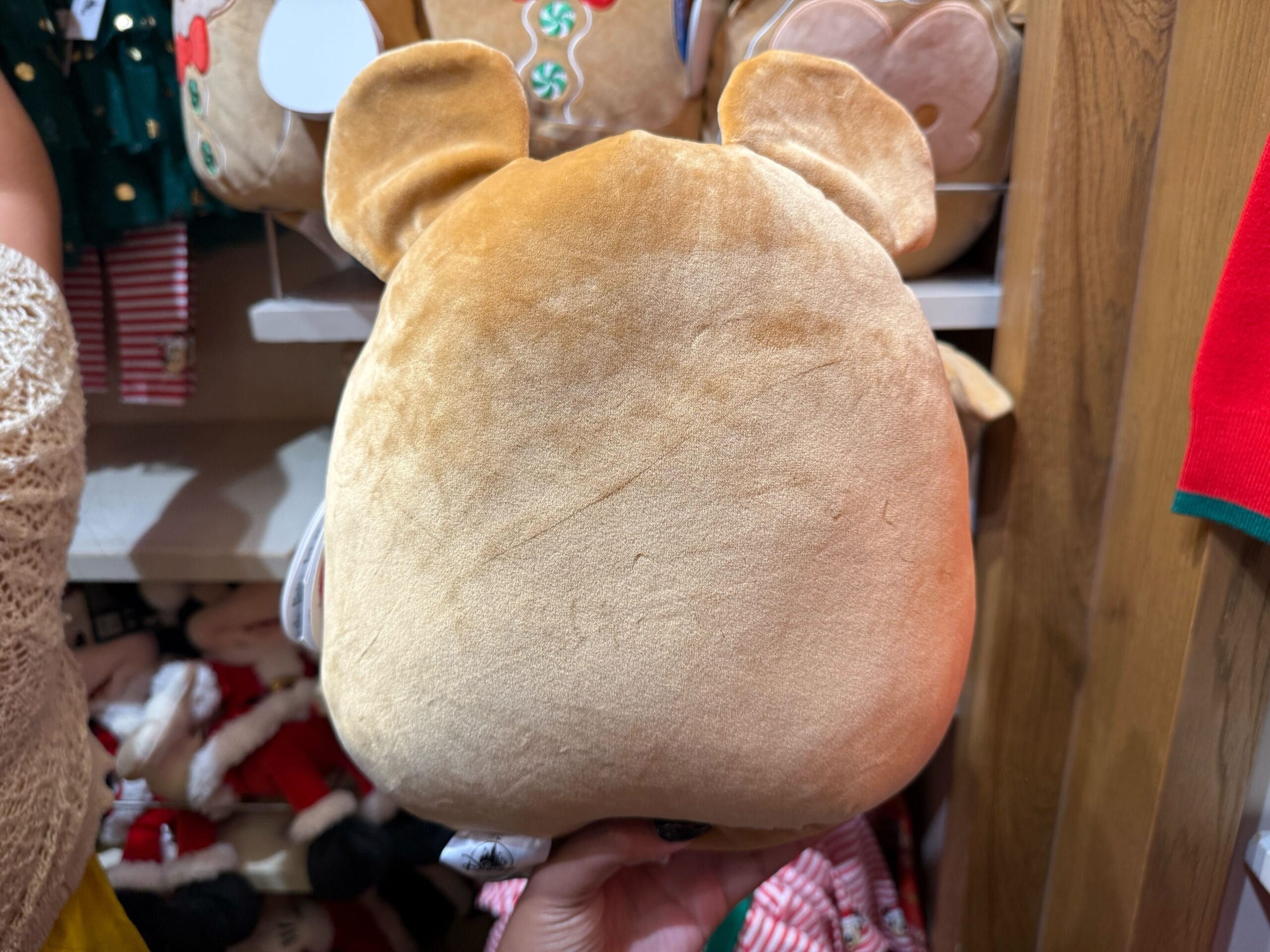 A guest holds a tan teddy bear plush by Disneyland Christmas merchandise, surrounded by plush toys in a Disney park shop.