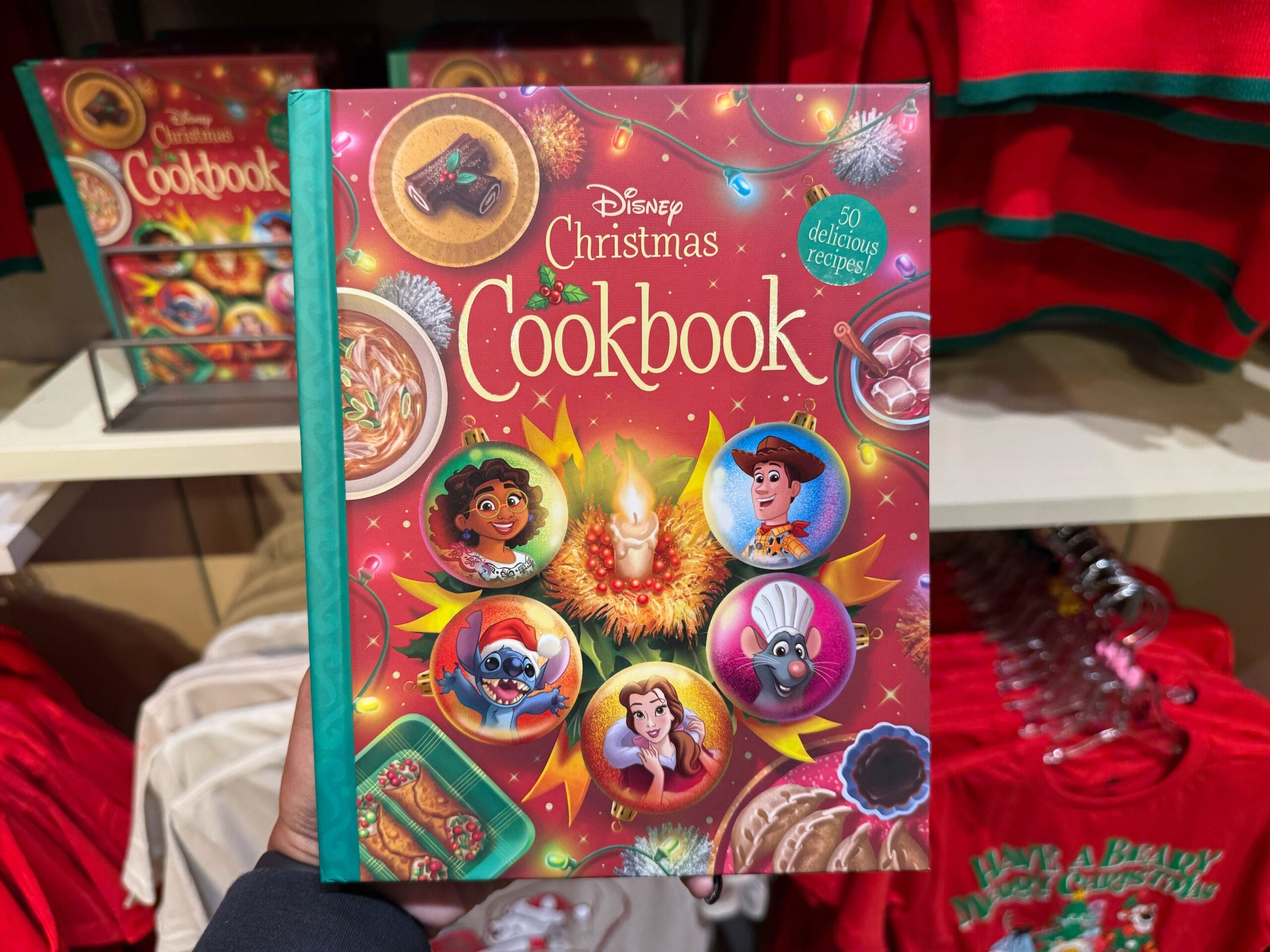 A hand holds a Disney Christmas Cookbook among festive shop displays, surrounded by Disneyland holiday merchandise—noting this resembles Main Street, with castle decor more like Disneyland California’s charming Sleeping Beauty Castle than the grander Cinderella Castle of Walt Disney World.
