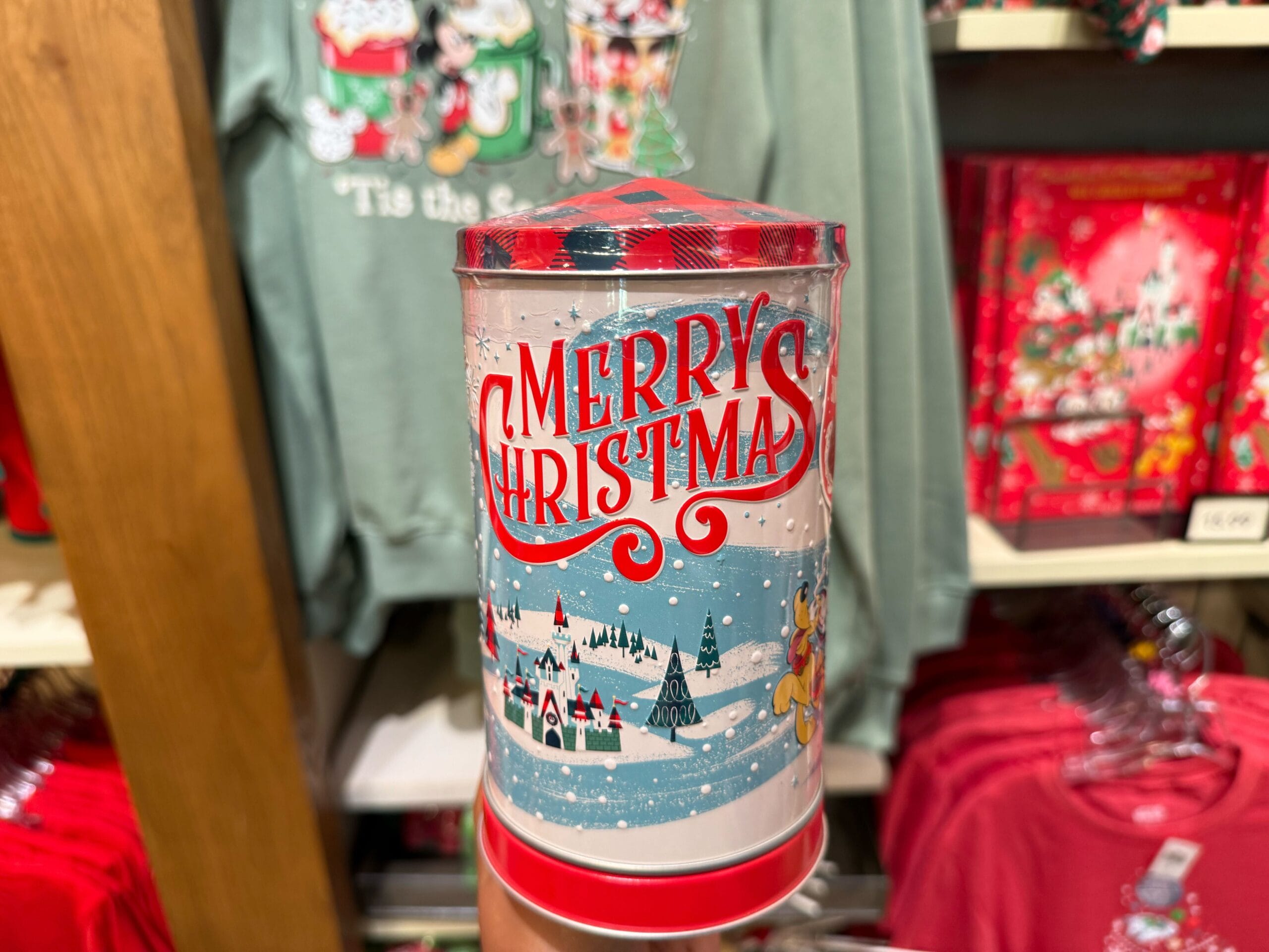 A hand holds a "Merry Christmas" tin with a snowy scene, Disneyland holiday merchandise behind—reminiscent of Main Street shop displays.