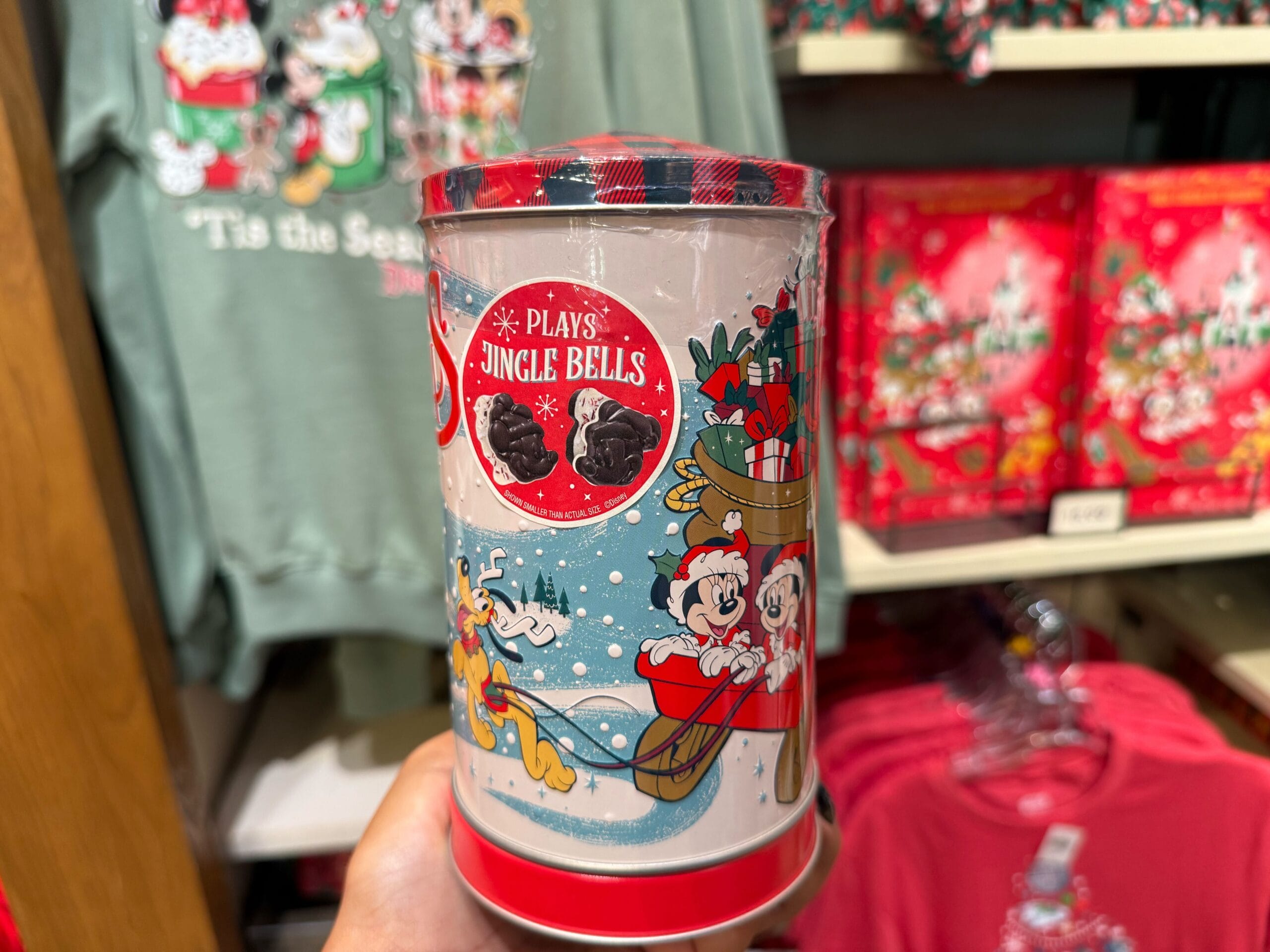 A hand holds a festive tin with cartoon mice in a sleigh; Disney Peppermint Collection shirts displayed behind, Main Street style.